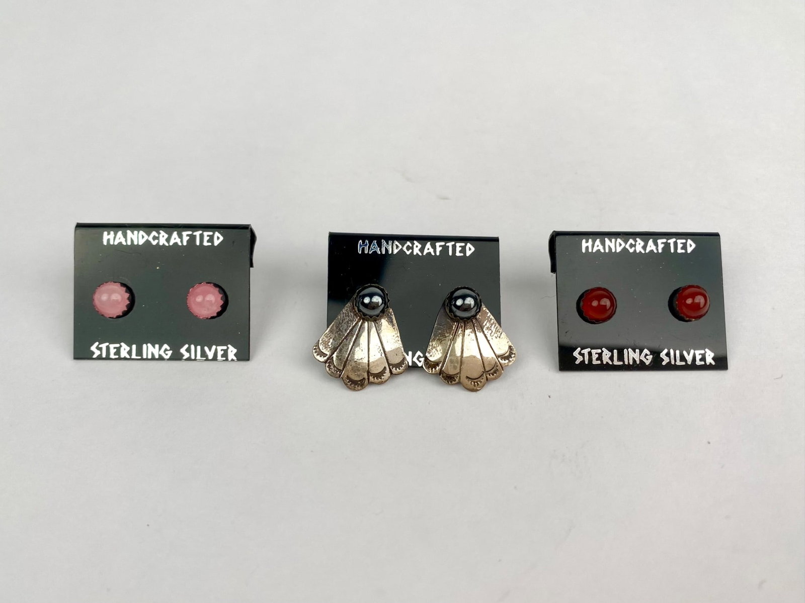Lot of Three Pairs of Handcrafted Sterling Silver Earrings on Original Cards: Set of three pairs of handcrafted sterling silver earrings. The first pair features pink circular cabochon stones, the second pair highlights fan shaped designs with metallic black accents, likely hem