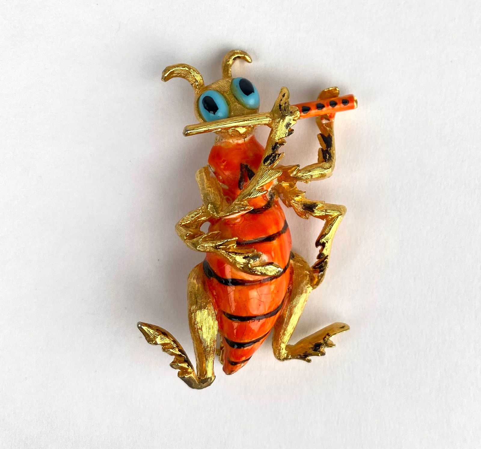 Hattie Carnegie Enamel and Gold Tone Figurative Insect Brooch: Hattie Carnegie insect brooch featuring vibrant orange enamel, gold tone metal detailing, and blue eyed accents. The insect is depicted playing a flute, adding a playful touch to the design. The back