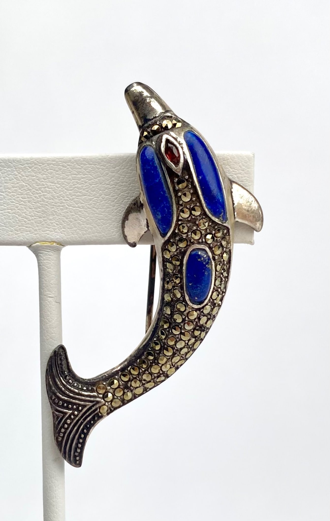 Sterling Silver Dolphin Marcasite Lapis Lazuli and Garnet Stone Detail: Sterling silver dolphin shaped brooch featuring Lapis Lazuli inlays on the body and a small Garnet stone detail as the eye. Accented with marcasite stones, adding texture and shine. The brooch has a w