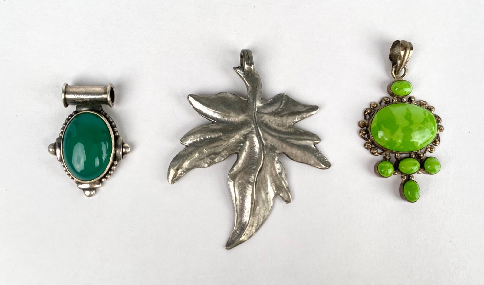 Set of Three Sterling Silver Pendants With Green Stone Accents: A collection of three sterling silver pendants. The first pendant features a green cabochon stone in an ornate setting, marked 925. The second pendant displays a green oval stone accompanied by smalle