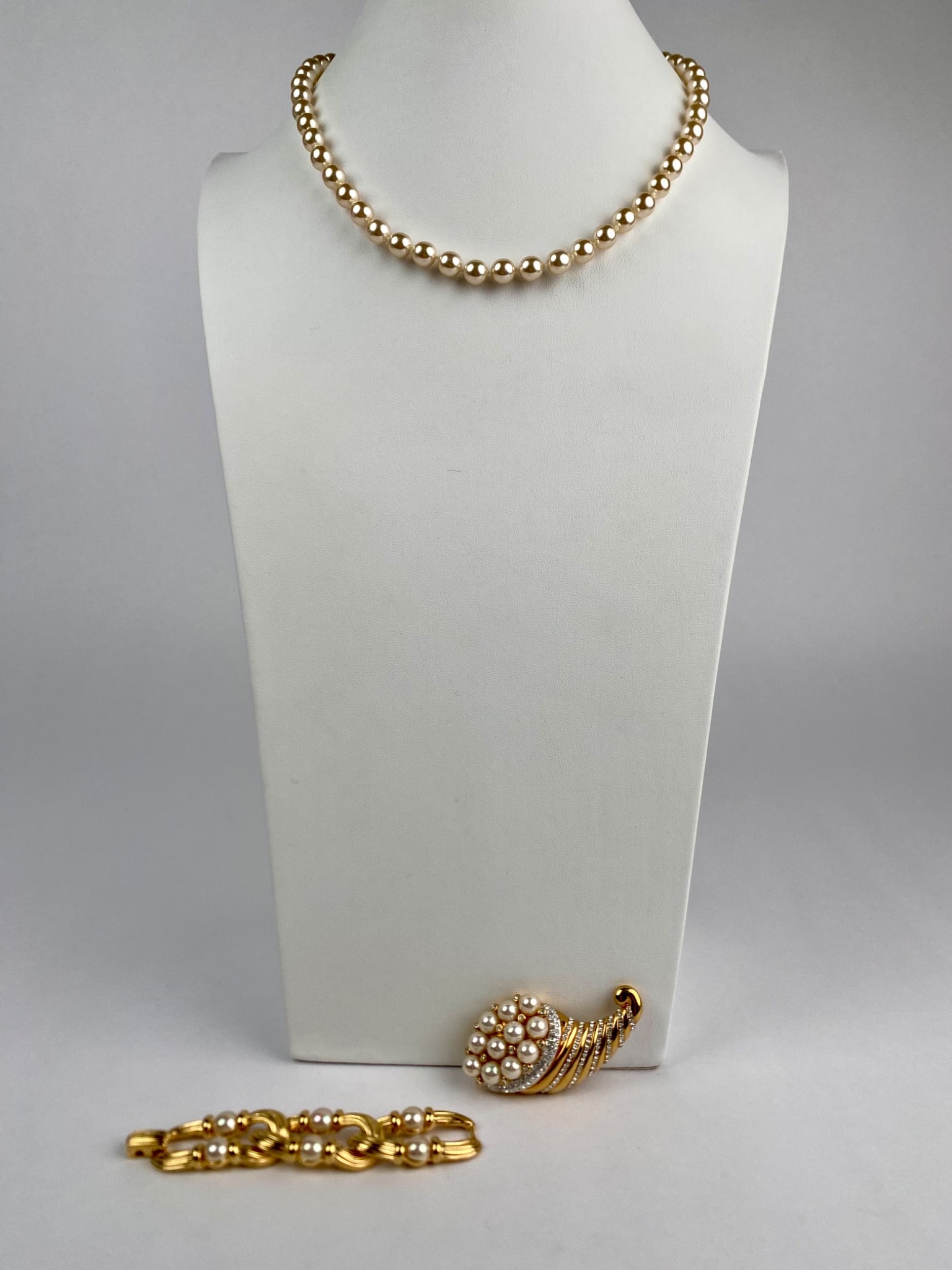 Nolan Miller Faux Pearl Necklace, Brooch, and Bracelet Link Set: Nolan Miller jewelry set featuring a gold tone faux pearl necklace with a screw clasp adorned by clear rhinestones, a brooch in a cornucopia design with faux pearls and rhinestones, and a gold tone an