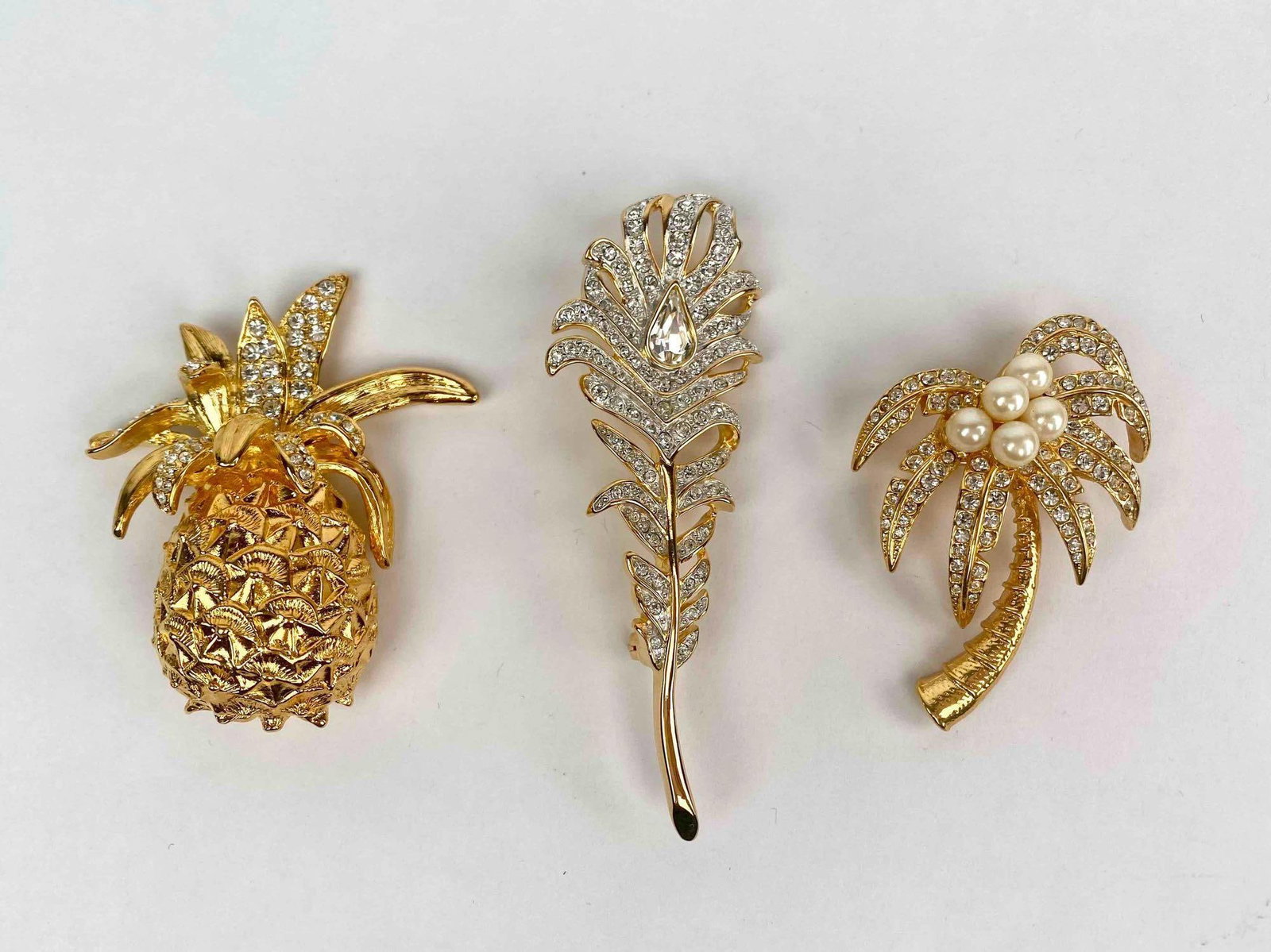 Lot of three signed Kenneth Jay Lane (KJL) brooches, each showcasing tropical inspired designs.: Lot of three signed Kenneth Jay Lane (KJL) brooches, each showcasing tropical inspired designs. Included in this lot are a rhinestone studded gold tone pineapple, a sparkling palm tree accented with f
