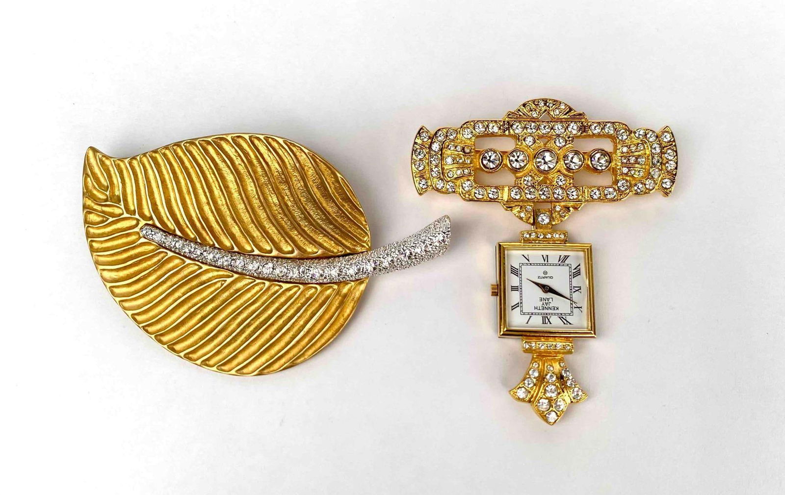 Pair of brooches by Kenneth Jay Lane. One brooch is a gold tone leave with a silver tone and: Pair of brooches by Kenneth Jay Lane. One brooch is a gold tone leave with a silver tone and rhinestone center. The second brooch is a hanging clock with rhinestone details. Both brooches are hallmark