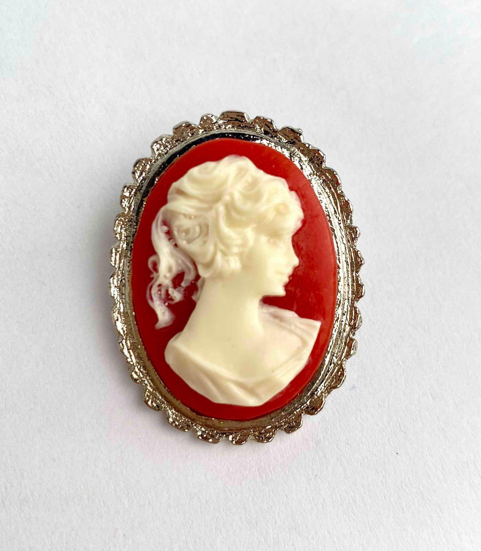 A vintage costume cameo brooch featuring a high relief white female bust set against a vibrant red: A vintage costume cameo brooch featuring a high relief white female bust set against a vibrant red background. Framed in an ornate scalloped silver tone setting. Brooch is in good condition. Please se