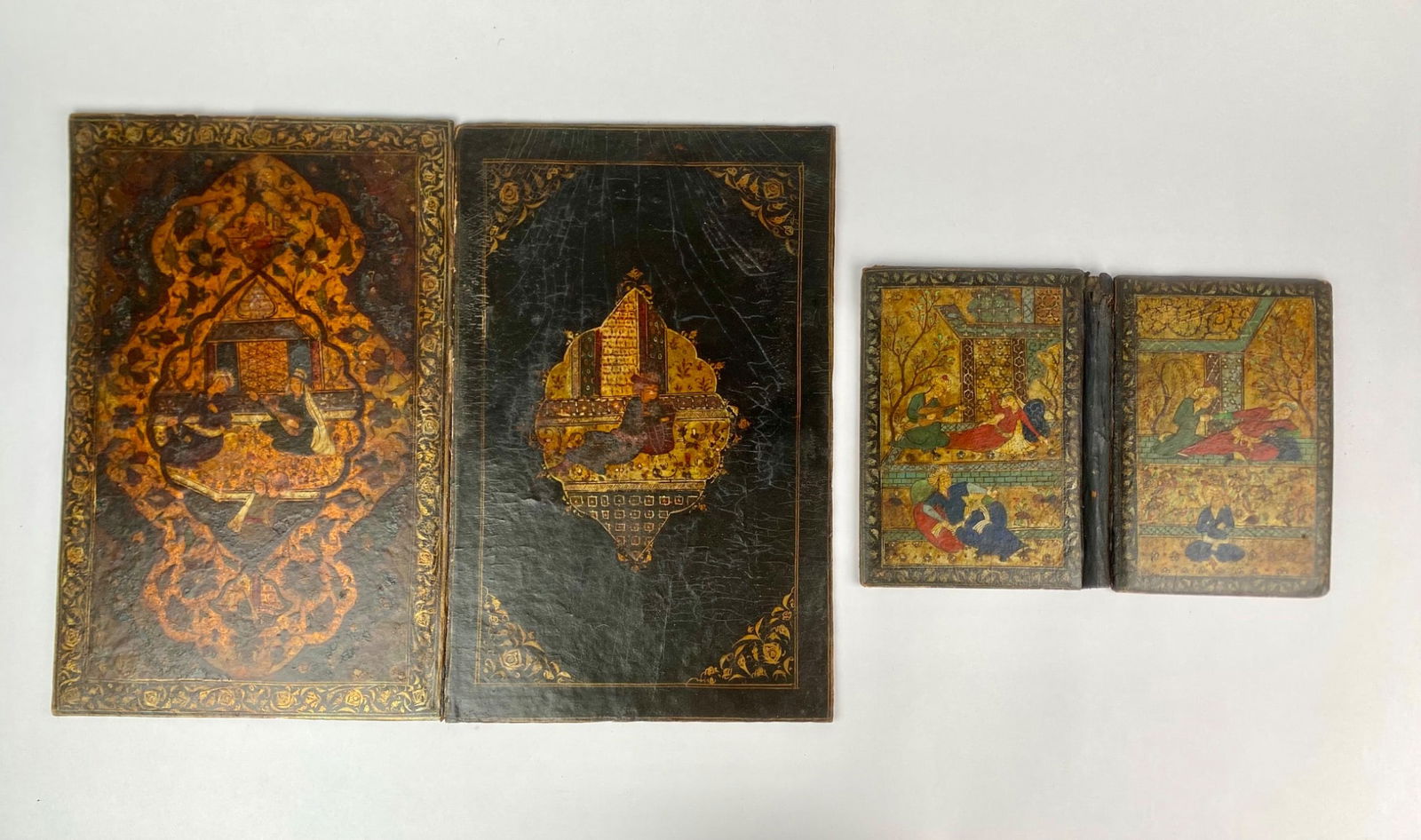 Four Persian Style Hand Painted Lacquered Book Covers: Set of four Persian or Persian style hand painted lacquered book covers, offered as decorative objects without interior pages. The two larger covers feature elaborate miniature scenes of poets and mus