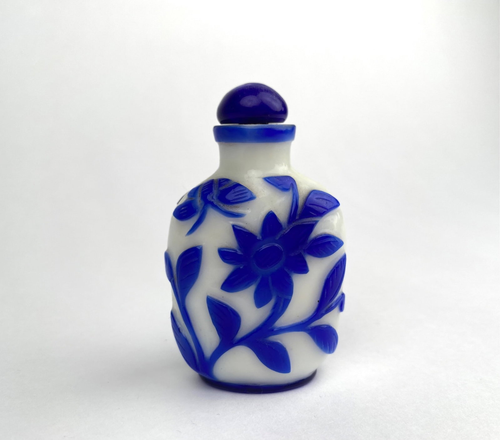 Chinese Peking Glass Overlay Snuff Bottle (Blue and White): Chinese peking glass blue and white overlay glass snuff bottle featuring a floral motif in relief, complete with a matching stopper. The bottle showcases intricate blue overlay against a white backgro