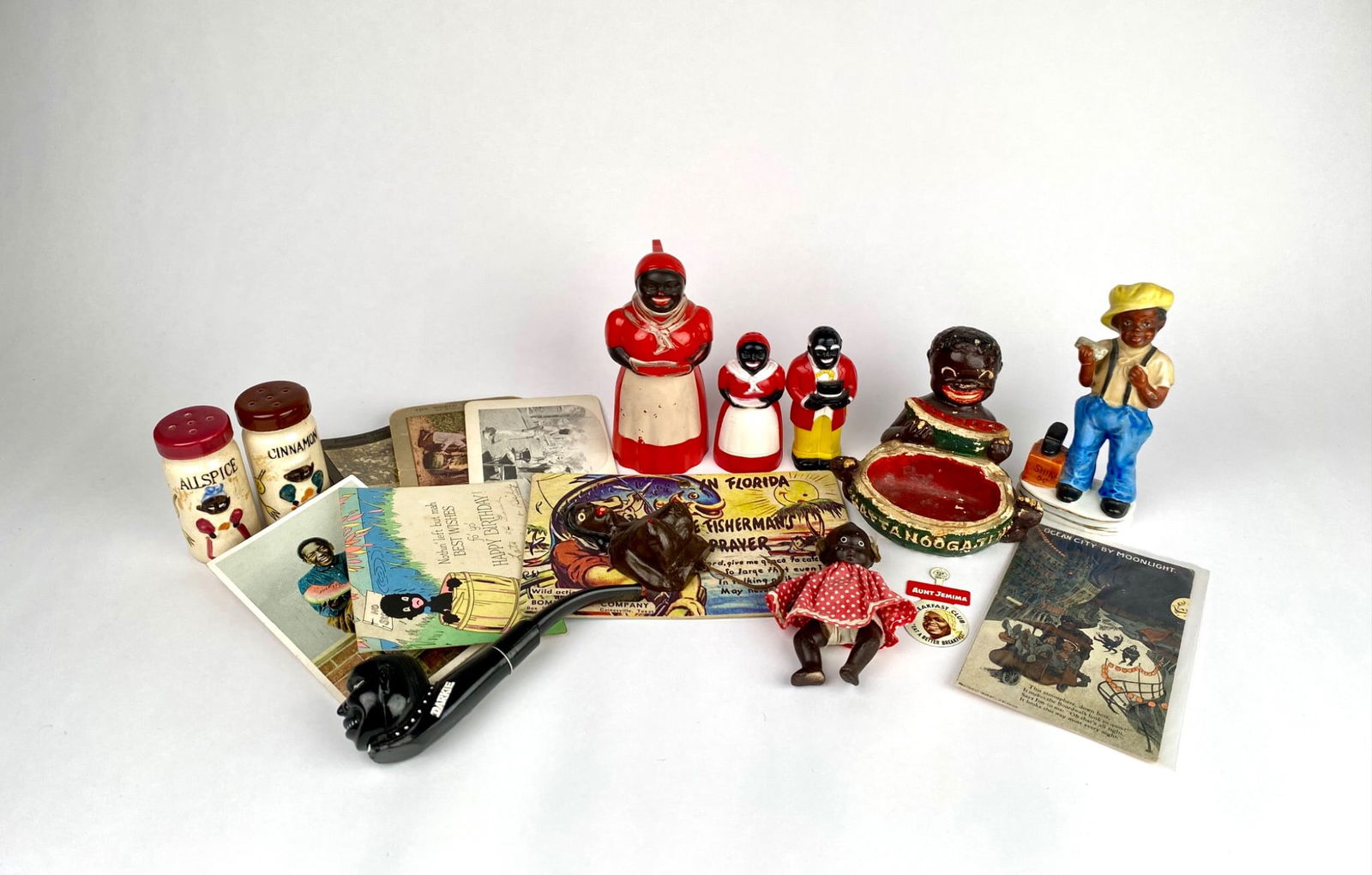 Lot of Black Americana Ephemera and Figurines: A diverse collection of Black Americana items, including vintage ceramic figurines, spice jars marked ALLSPICE and CINNAMON, a pipe labeled DARKIE, postcards, stereoviews, and a small doll in a polka