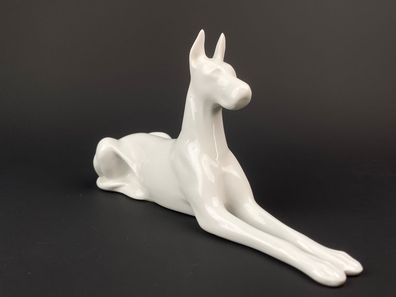 Porcelain Figurine of a Reclining Dog: This porcelain figurine is made with detailed craftsmanship. It shows a greyhound style dog in a resting position with a smooth white porcelain finish. The figurine highlights the dog's slender limbs,