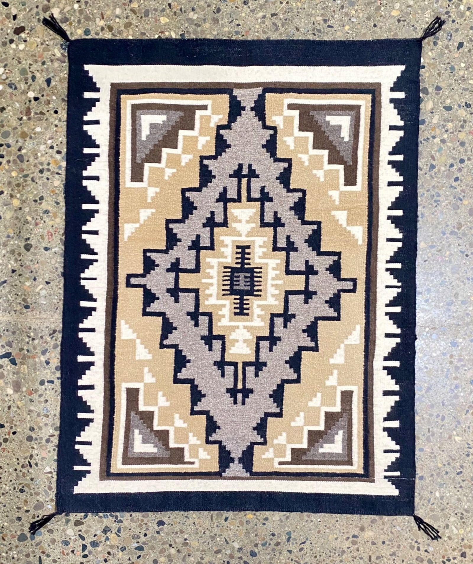 Two Grey Hills Navajo Rug: Handwoven rug from Two Grey Hills featuring an intricate geometric pattern in shades of beige, brown, white, and navy blue. Made from wool, the rug incorporates traditional motifs and tasseled corners