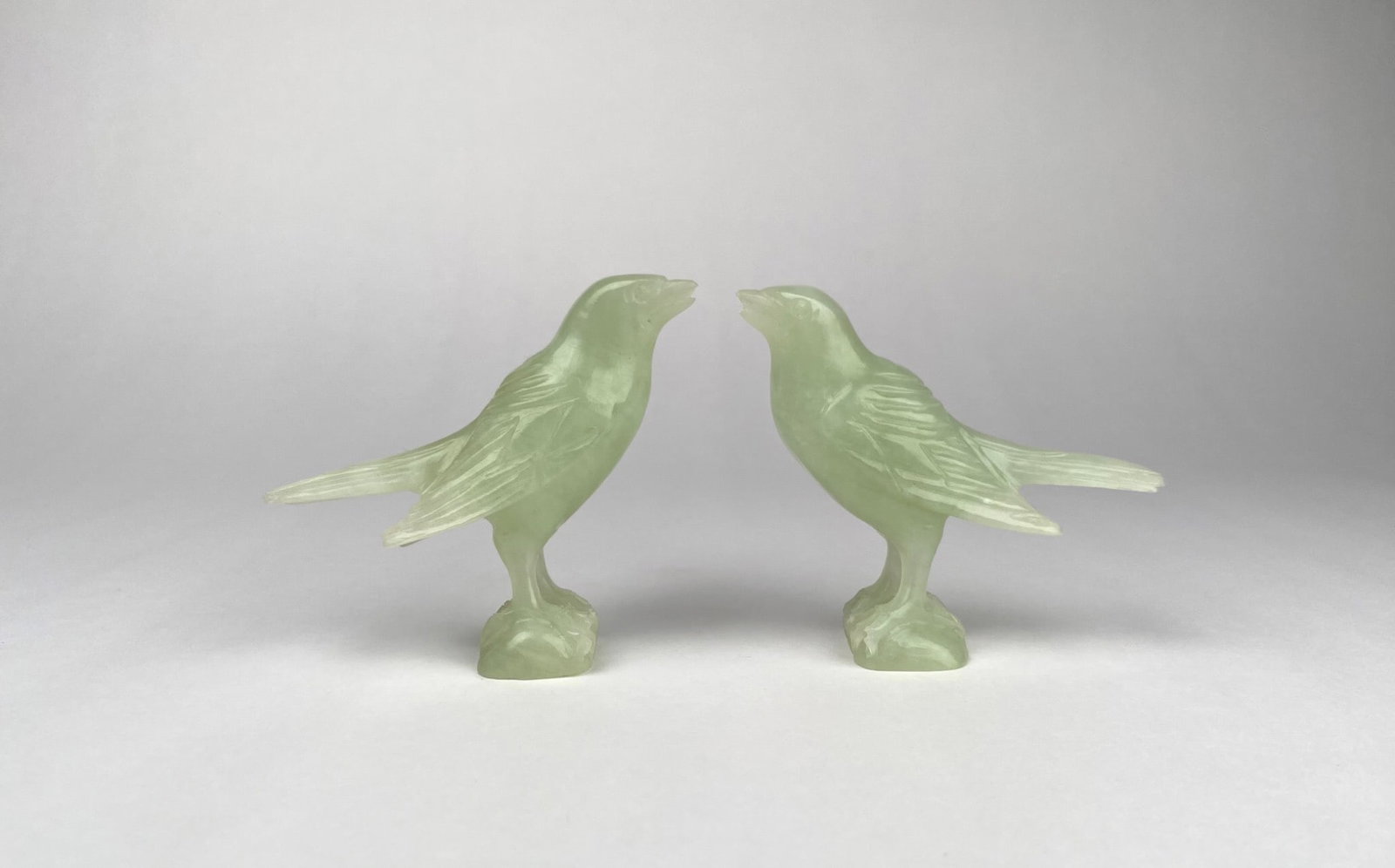 Pair of Carved Green Stone Bird Figurines: Green stone bird sculptures, each intricately carved with detailed feathers and poised on a naturalistic base. The sculptures exhibit a delicate green hue. Both birds are in good condition. Please see