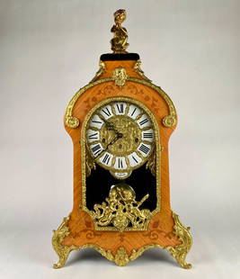 Italian Rococo Mantle Clock with Franz Hermle Movement (Tiffany): Tiffany style mantel clock featuring finely crafted wood veneer with intricate inlays and gilded brass accents. The clock is adorned with decorative cherubic figures and an ornate finial. The dial has