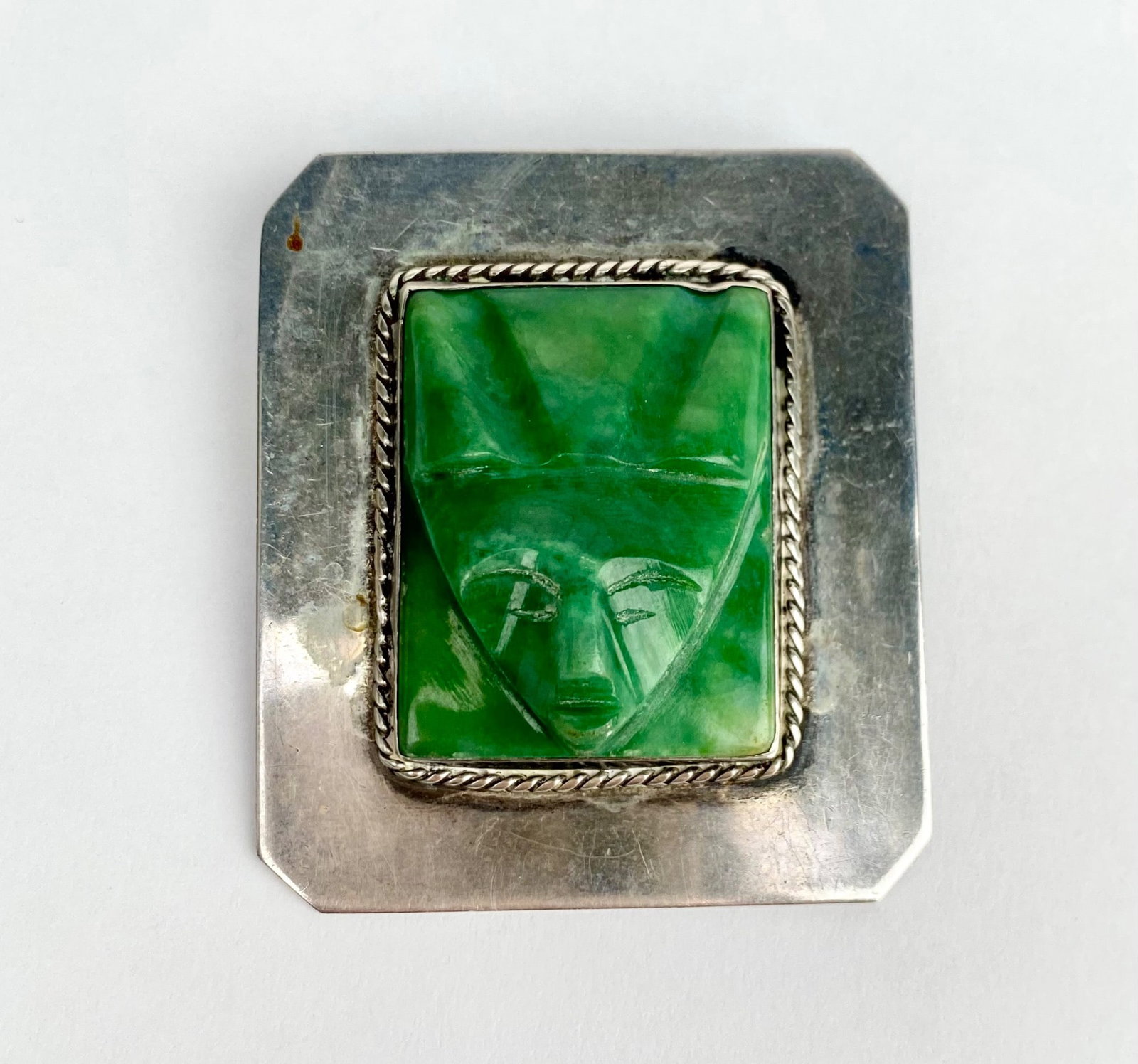 Mexican Sterling Silver Brooch with Green Face Carving: Mexican sterling silver brooch featuring a carved green stone face centerpiece accented with a rope style silver border. The piece is stamped SILVER MEXICO on the back. Includes a secure pin and clasp