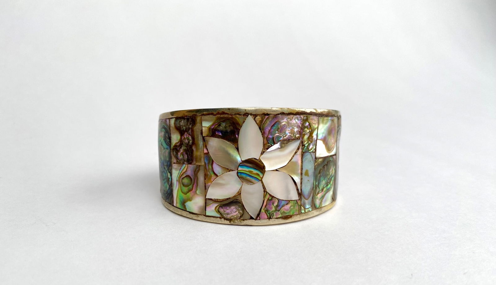 Abalone and Mother of Pearl Inlay Cuff Bracelet with Floral Design: Abalone and mother of pearl cuff bracelet featuring an inlaid floral design. The inlays display vibrant hues of green, purple, and blue against a metallic cuff. The inner band exhibits visible marking