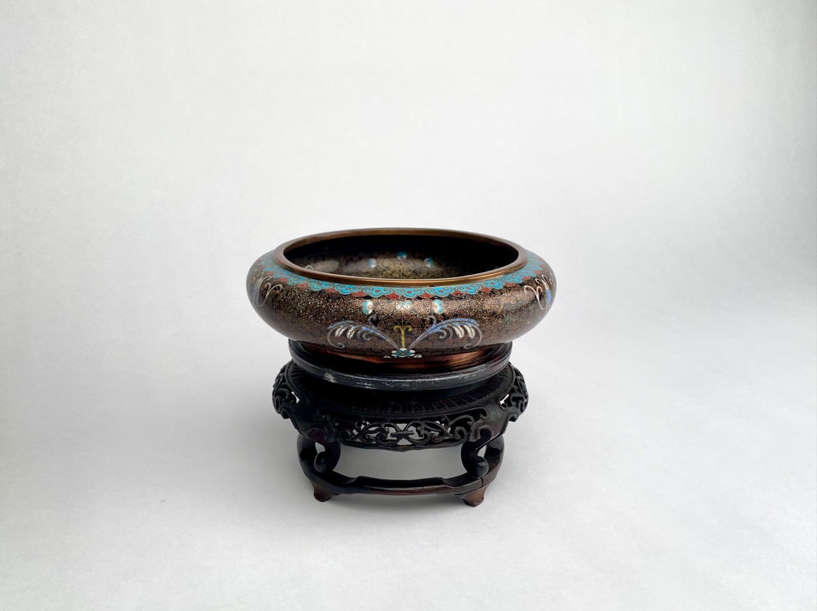 Chinese Cloisonne Enamel Bowl with Wood Stand: A Chinese cloisonne enamel bowl featuring intricate multicolor floral and geometric designs against a black background. The interior and base have sky blue enamel with a swirl pattern. The rim and edg