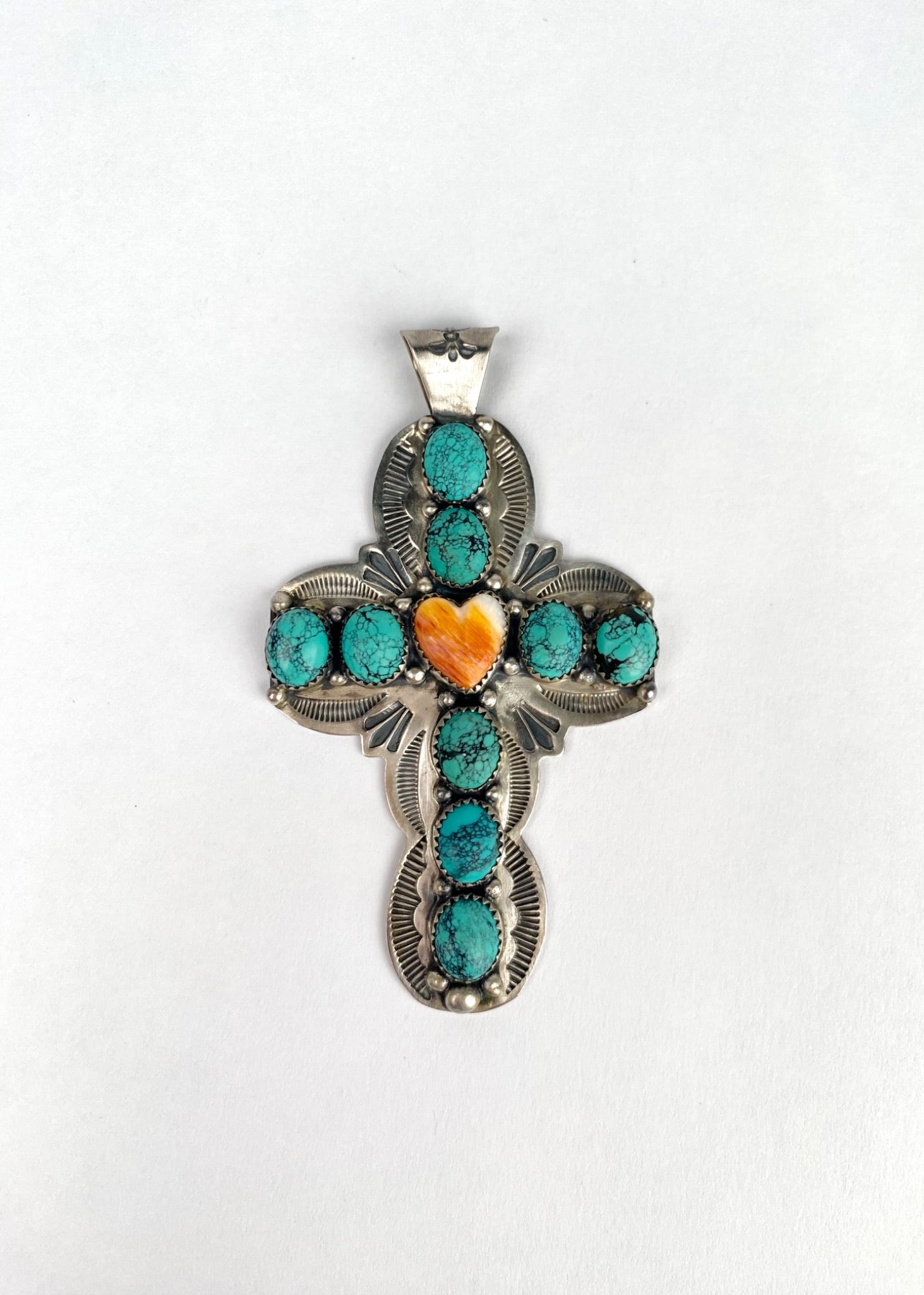 Navajo Silver Cross Pendant With Turquoise and Orange Stone Heart: Navajo sterling silver cross pendant featuring nine turquoise stones and one orange stone in the shape of a heart set amidst intricate silverwork. The back of the pendant bears a stamped mark indicati