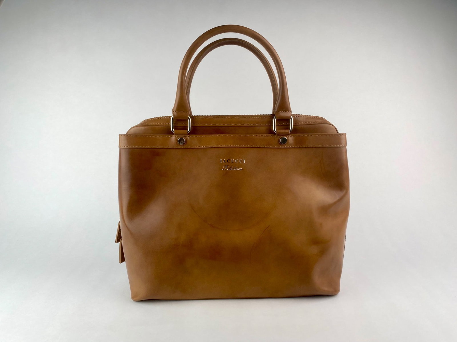 IACUCCI Italian Tan Leather Structured Handbag with Strap: Structured leather handbag by IACUCCI, crafted in tan genuine leather with dual rolled handles and silver tone hardware. The bag features a zip top closure, protective metal feet on the base, and a cl