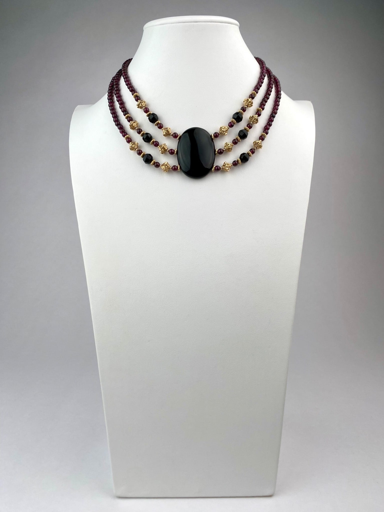Three-Strand Garnet and Onyx Necklace with Gold Tone Accents: Three-strand necklace featuring graduated deep red garnet beads strung in three rows, accompanied by faceted onyx beads and gold-tone spacers. A large, polished oval onyx cabochon acts as the centerpi