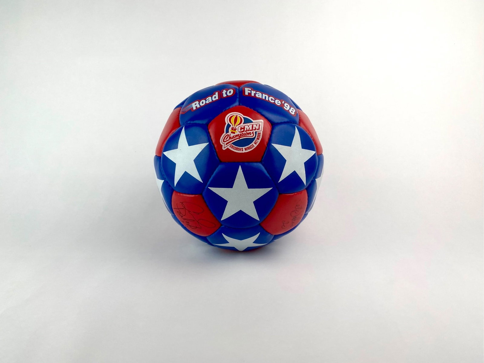 1998 Road to France USA Soccer Ball Signed by Danielle Fotopoulos, Kate Sobrero, and Cindy Parlow: Red, white, and blue soccer ball emblazoned with stars and promotional text reading "Road to France '98," commemorating the 1998 FIFA World Cup campaign. The ball features logos from sponsors AMOCO an