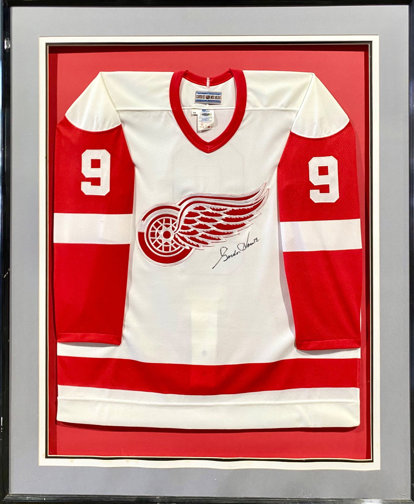 Framed Gordie Howe Autographed Detroit Red Wings Hockey Jersey: Framed Gordie Howe Detroit Red Wings hockey jersey with a red and white color scheme, featuring the iconic winged wheel logo and the number 9 on the sleeves. The jersey bears a signature in black ink.