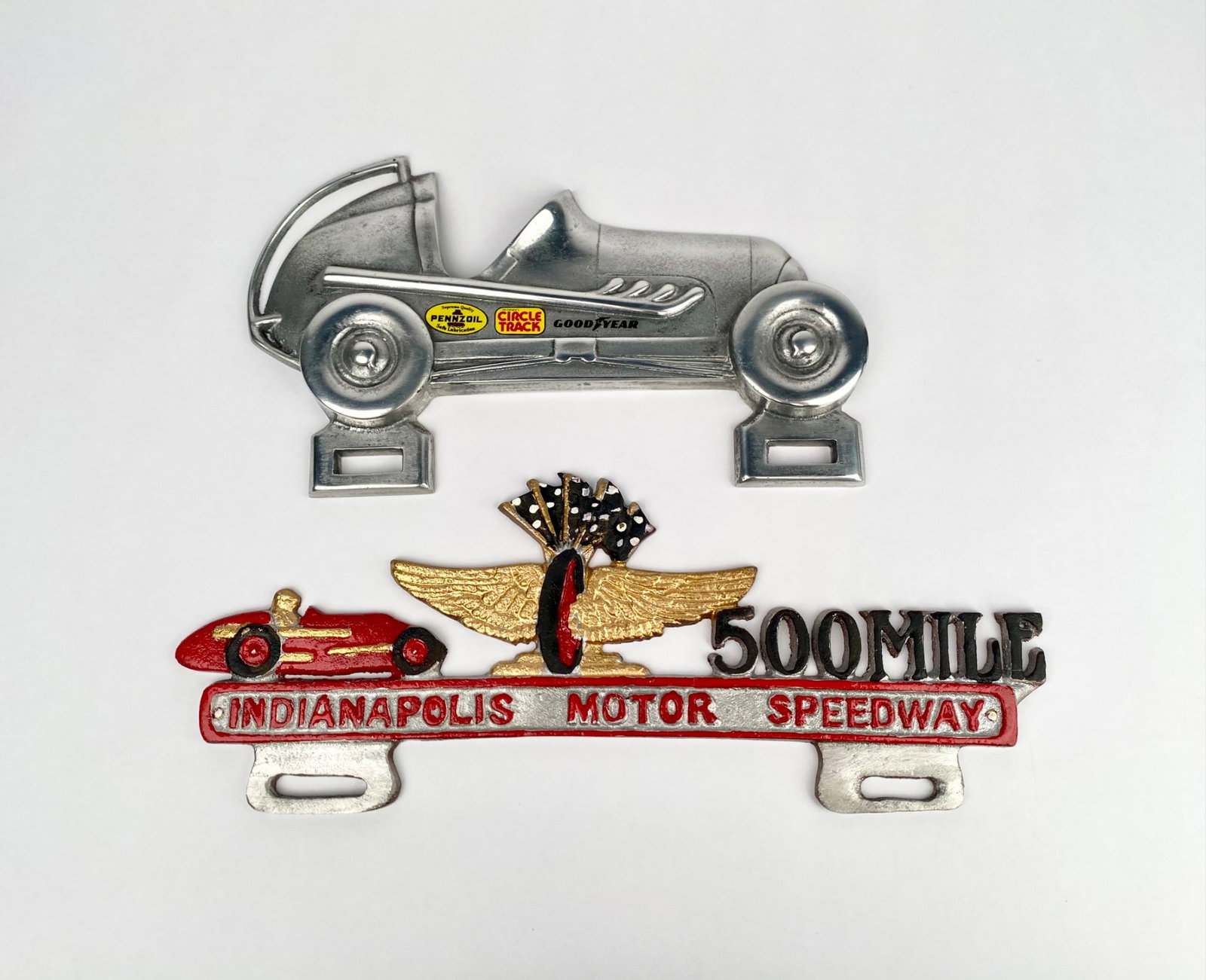Two Indianapolis Motor Speedway Themed Metal License Plate Toppers: Two vintage-style metal license plate toppers featuring Indianapolis Motor Speedway themes. One is a 500 Mile motif with gold and red enamel paint detailing, and the other depicts a silver racing car 