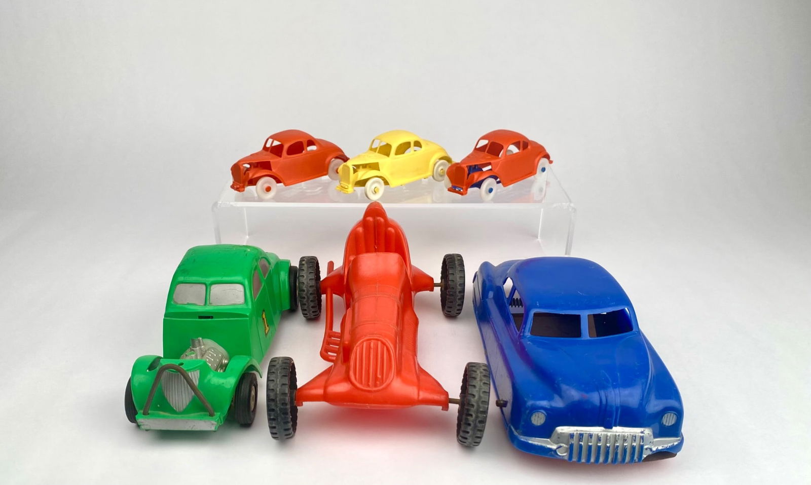 6 Vintage Plastic and Metal Toy Cars in Assorted Styles and Colors: Six toy cars, made of plastic and metal, featuring varied styles, including three smaller cars in orange, yellow, and red with molded numbers on top (49, 67, 85), one green coupe with a detailed metal