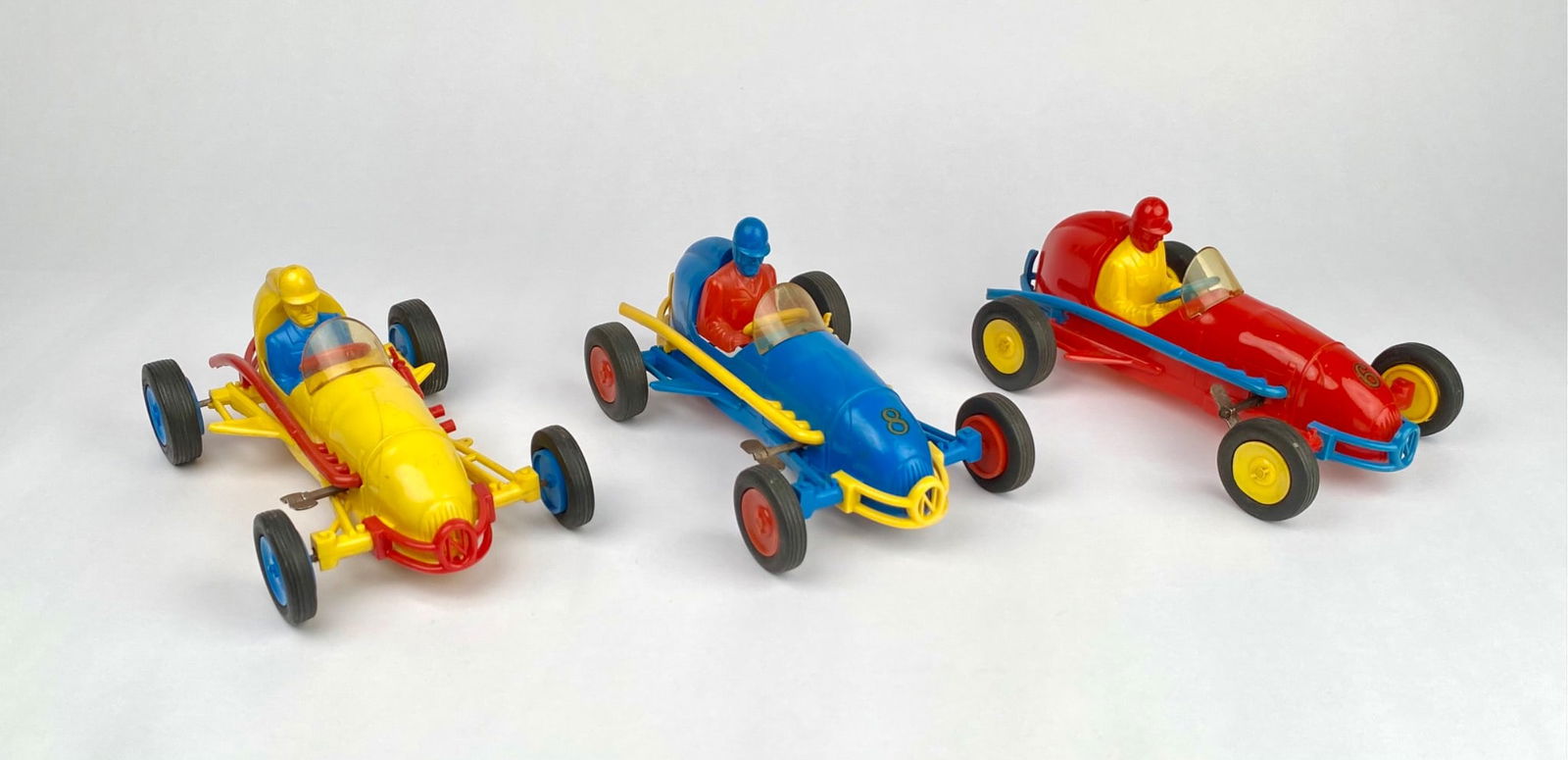 Lot of 3 Vintage Auburn Rubber Toy Indy Race Cars: Three vintage Auburn Rubber toy race cars in yellow, blue, and red. Each car features a driver figure, rubber wheels, and windshield. The cars are equipped with metal weight components mounted on the