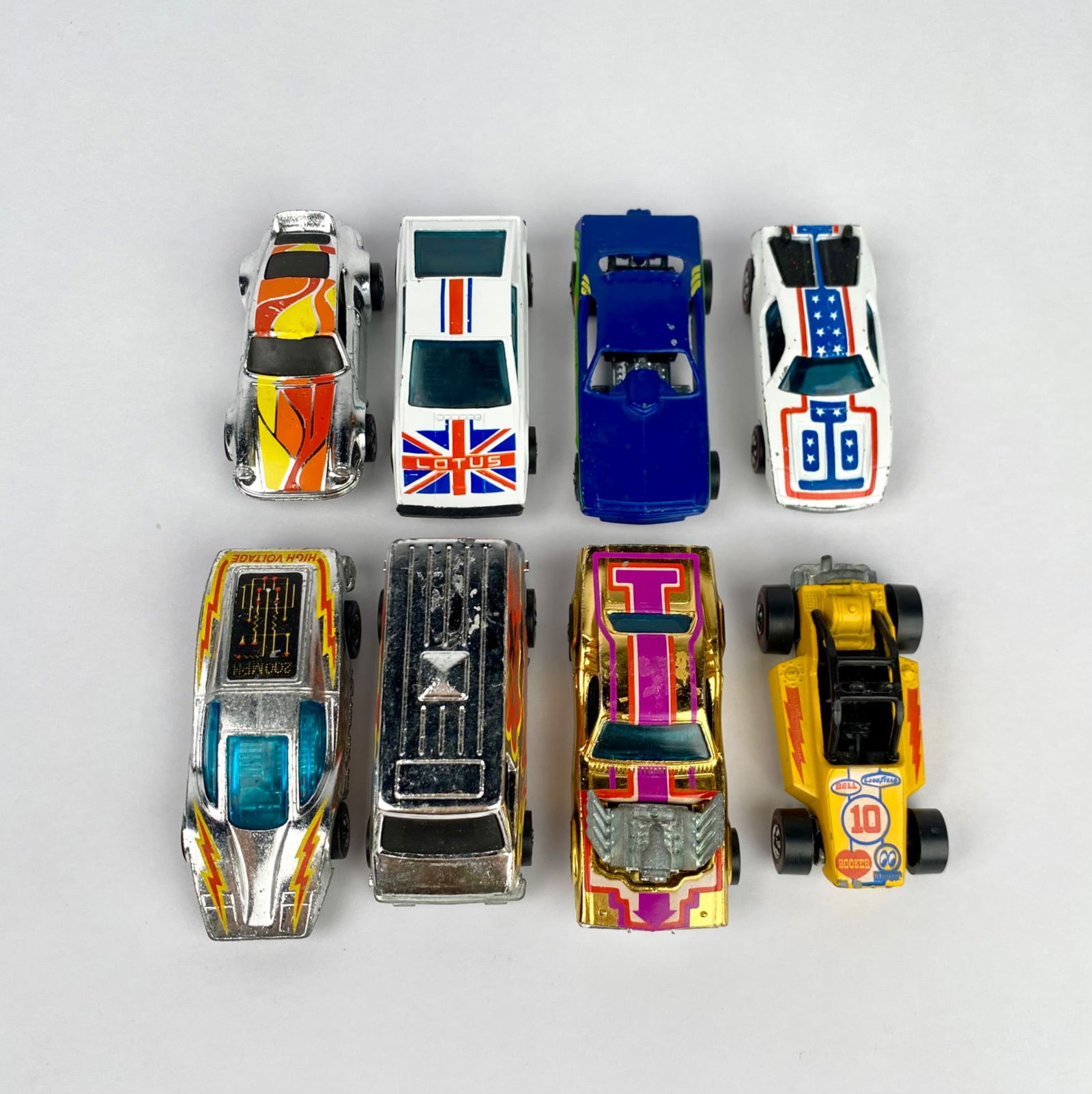 8 Vintage Hot Wheels Cars, 1970s-1980s: Eight vintage Hot Wheels die-cast toy cars featuring varied designs, including colorful decals such as a Union Jack, racing stripes, and flame motifs. Markings on the undersides indicate Hot Wheels br