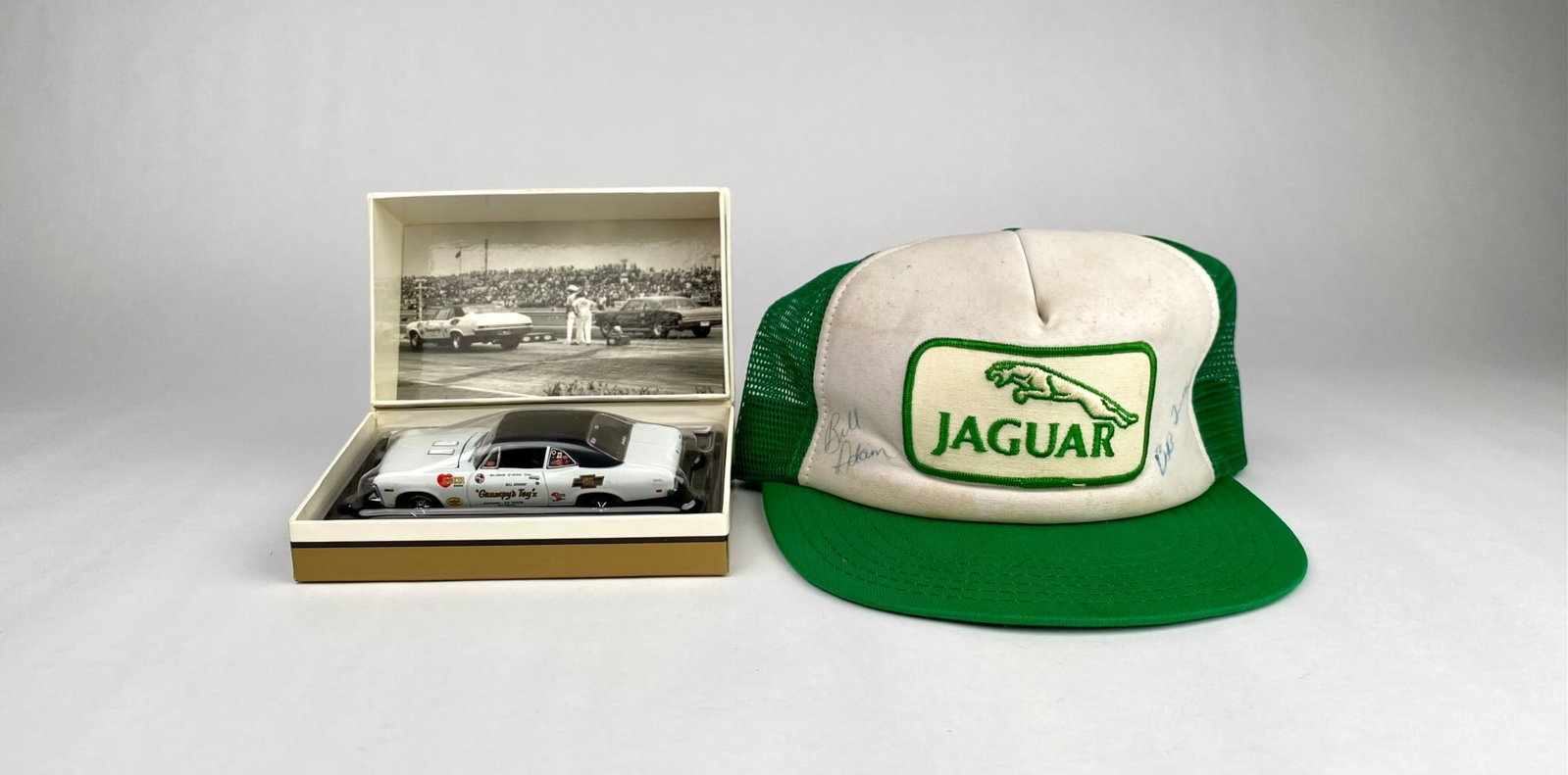 Bill 'Grumpy' Jenkins Signed Jaguar Racing Hat with GMP Diecast: A green and white Jaguar trucker hat featuring autographs and showing wear typical of age and use, alongside a GMP scale model of the 1968 Bill 'Grumpy' Jenkins Chevrolet Nova SS396. The model is pres