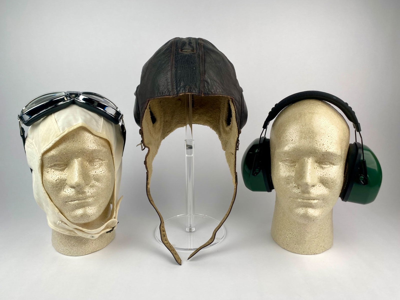 Set of Three Headgear- WWII Leather Flight Helmet, Canvas Cap w/ Goggles and Field & Stream Earmuffs: Lot includes a World War II-era leather flight helmet with shearling lining, twin receiver pockets/ear cups, chin strap, and goggle-strap loops. Also included is a white canvas racing-style cap paired