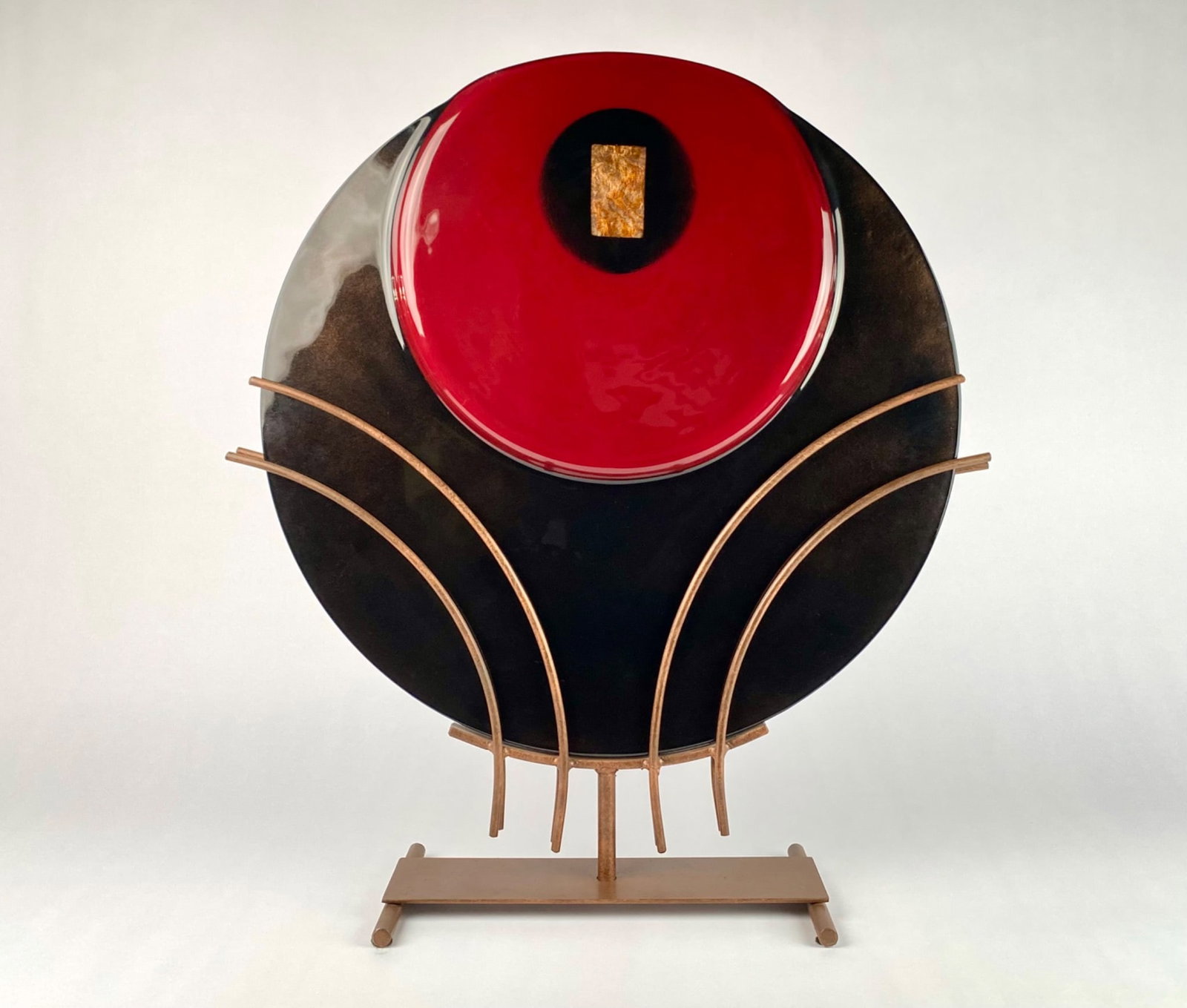 Matt Hines Glass and Metal Sculpture: Glass and metal sculpture by Matt Hines featuring a striking red and black circular glass piece accented by a central golden rectangular detail. Mounted on an intricate golden metal frame and stand. S