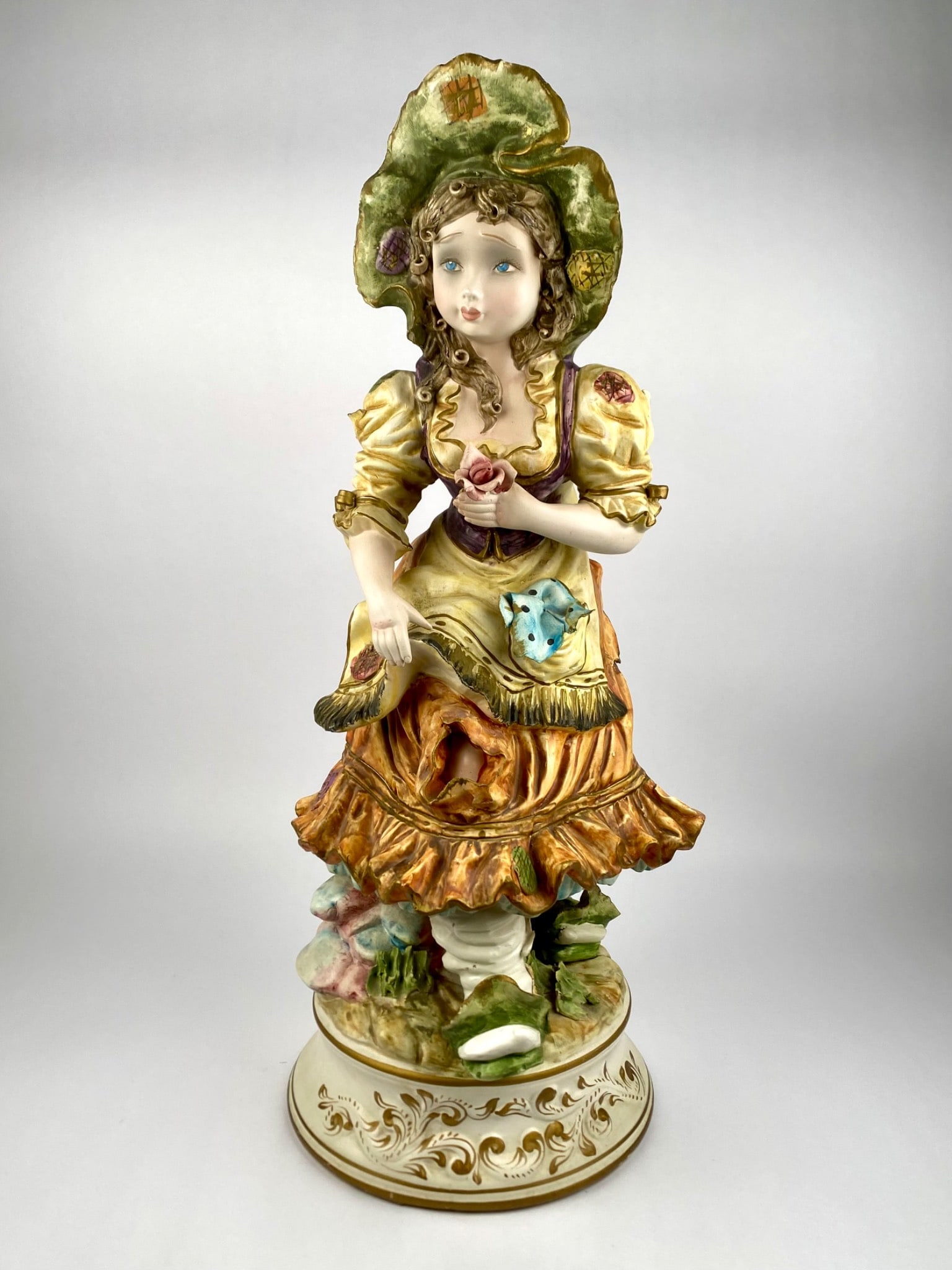 Italian Capodimonte-Style Hand-Painted Porcelain Figurine of Girl with Bonnet and Rose: Large Italian Capodimonte-style porcelain figurine depicting a young girl with a rose in hand, wearing a wide green bonnet and ruffled yellow dress accented with painted patchwork motifs. The piece is