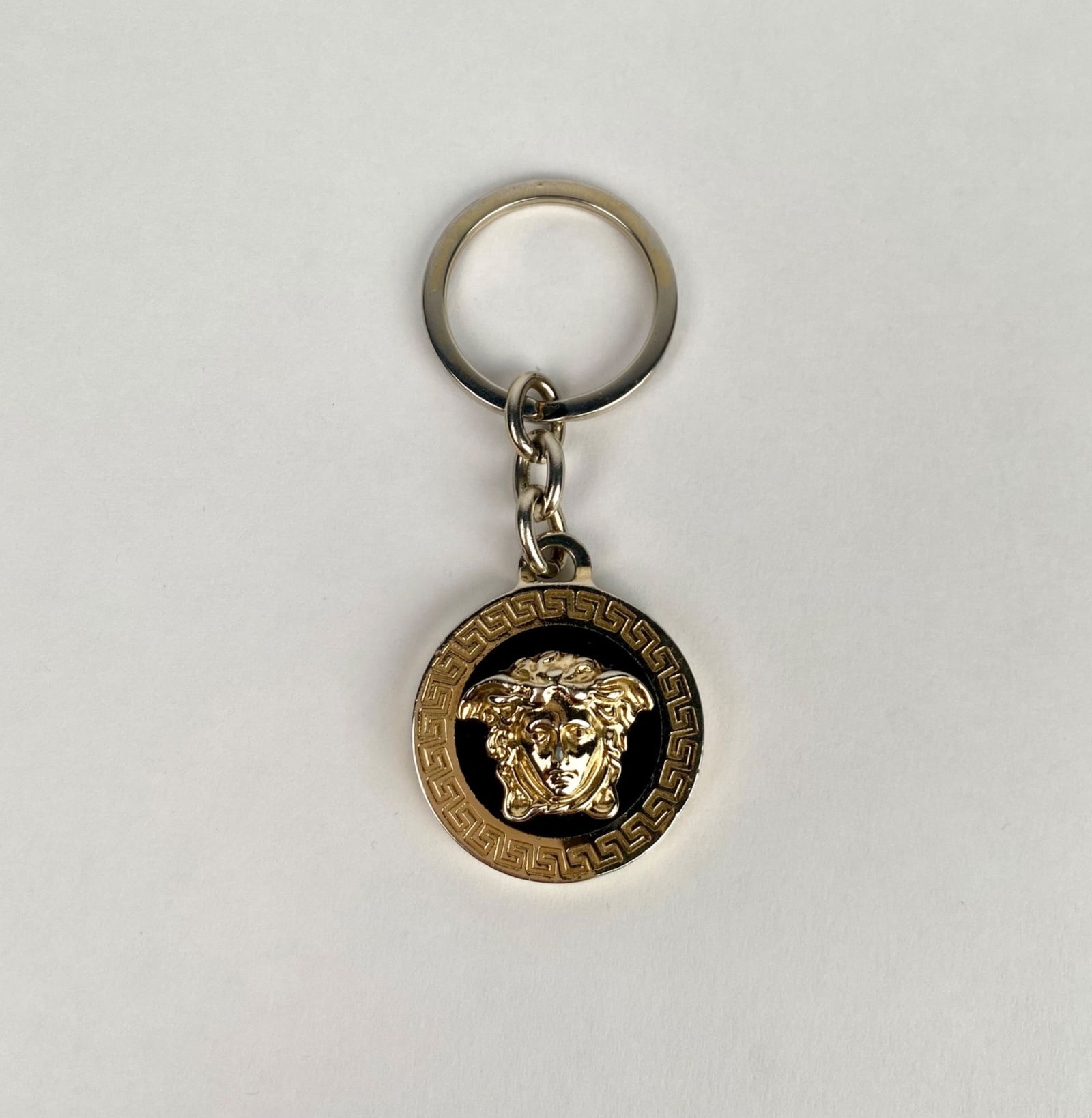 Versace Medusa Head Keychain: Gold tone Versace keychain featuring a Medusa head design against a black enamel background, framed by a Greek key border. The rear side is inscribed with VERSACE MADE IN ITALY. Includes a standard ke