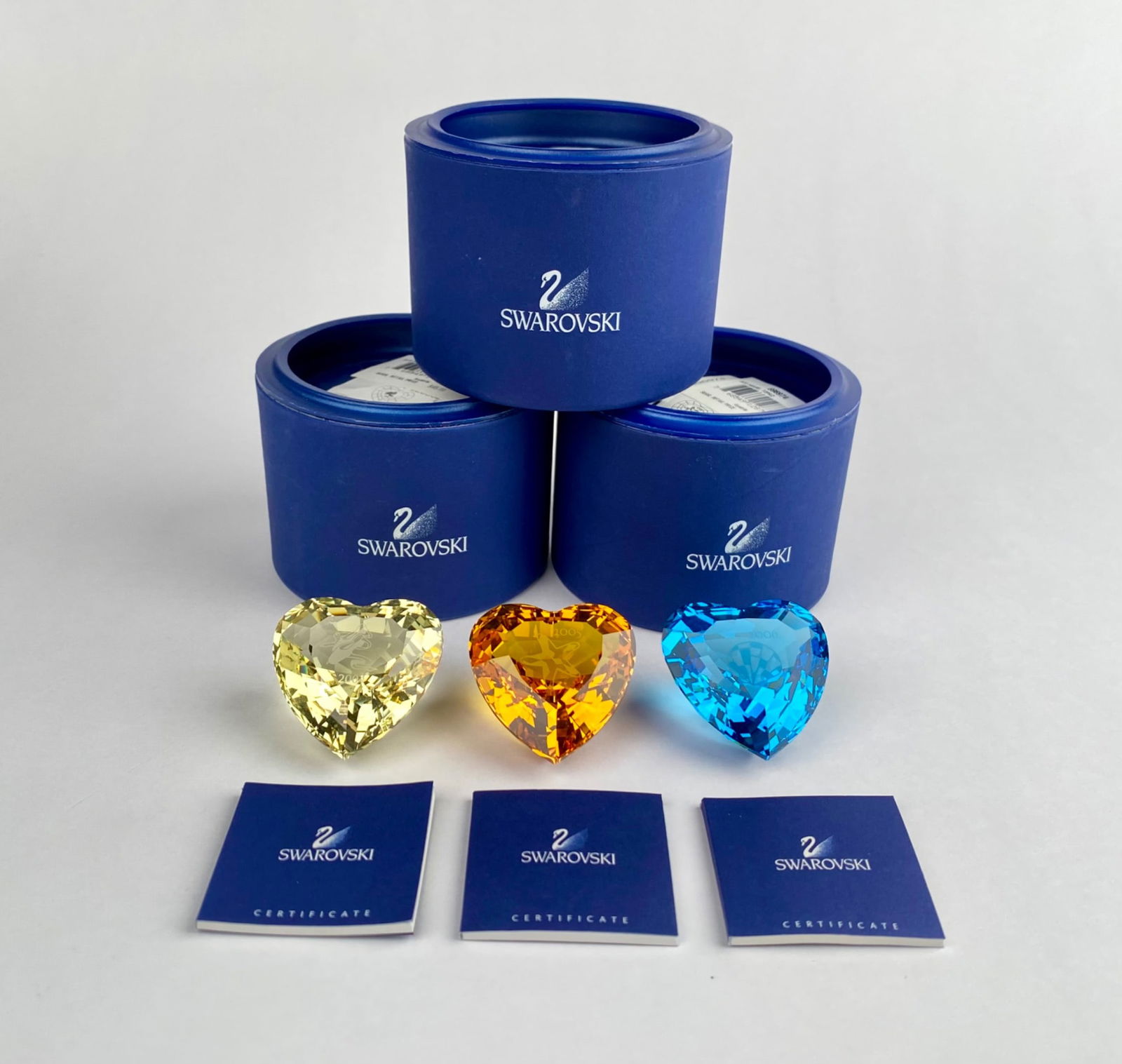 Swarovski Crystal Heart Trio with Certificates and Original Boxes (2004-2006): Three Swarovski crystal hearts in yellow, orange, and blue, each engraved with ‘2004’, '2005', and '2006' crafted to a faceted heart shape. Comes complete with certificates of authenticity and ori