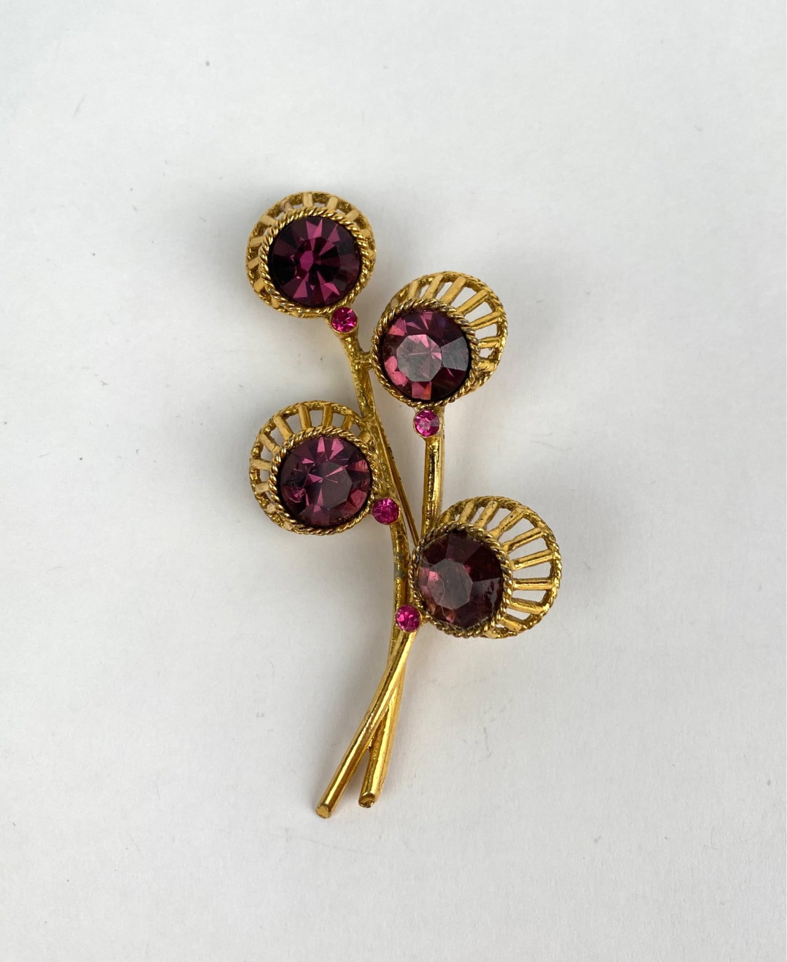 Mid Century Gold Tone Floral Brooch with Purple Rhinestones: Gold-tone floral brooch featuring purple rhinestones as flower accents. The intricate design includes openwork petals and smaller pink rhinestone details. The brooch shows craftsmanship in its metalwo