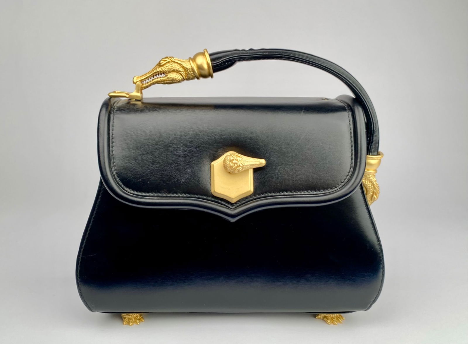 Black Leather Handbag with Gold Tone Ornamental Details: Black leather handbag featuring gold-tone hardware, including intricately designed crocodile-head handle attachments, paw motif feet, and a clasp with a matching crocodile motif. Interior lined with g