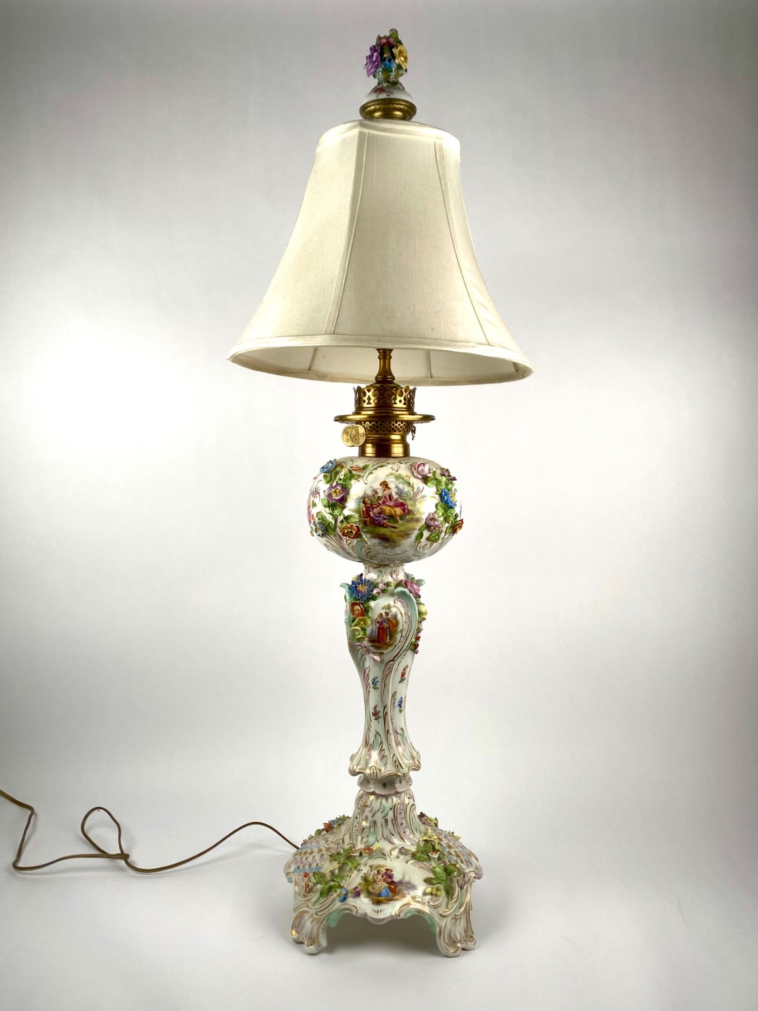 Large Dresden-Style Porcelain Figural Lamp with Hand-Painted Scenes and Applied Flowers: Large Dresden-style porcelain table lamp featuring hand-painted romantic courting scene in the Rococo style, framed by gilt accents and applied floral decorations throughout. The lamp is mounted with
