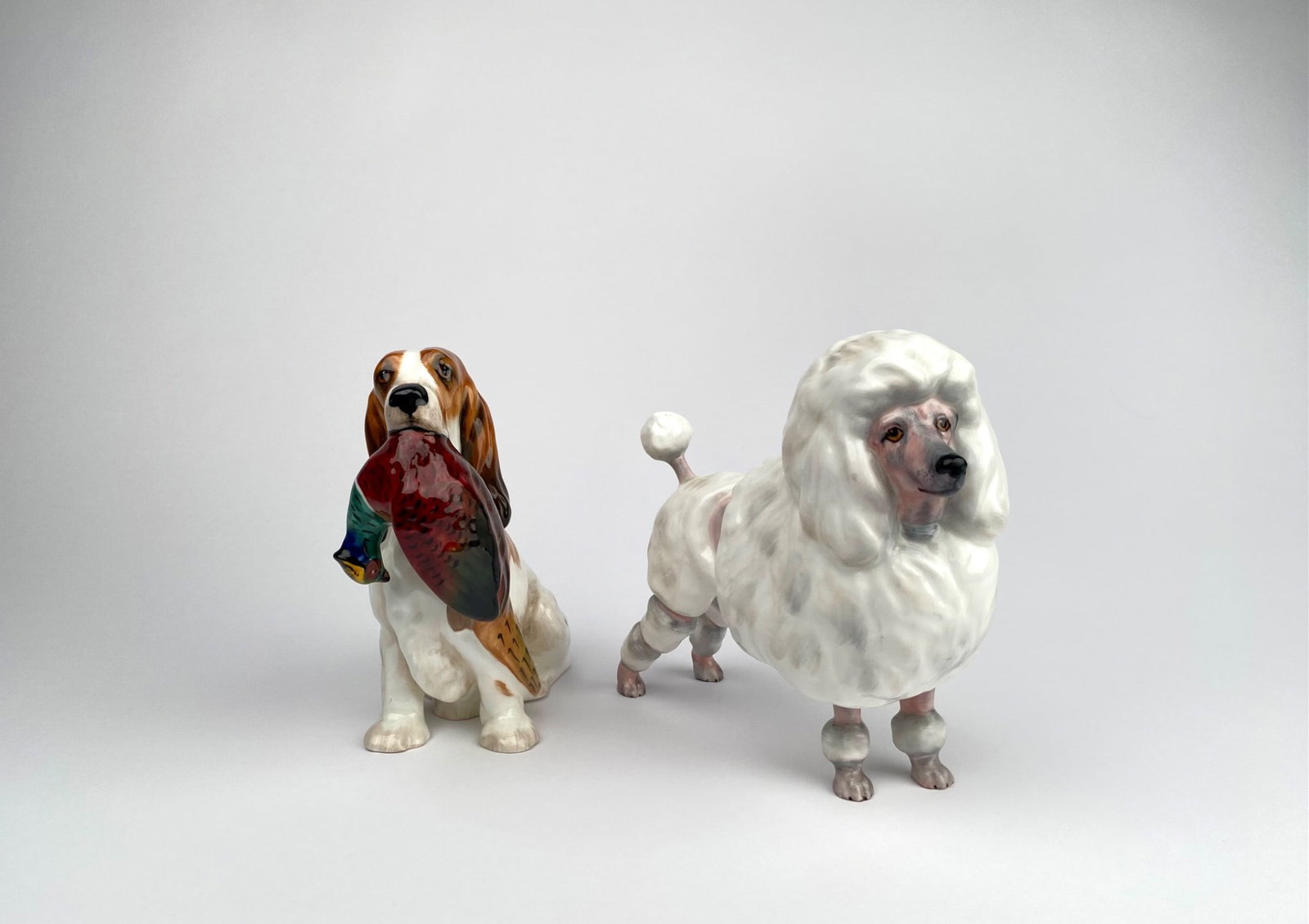 Royal Doulton Porcelain Figures of Poodle & Spaniel: Two Royal Doulton porcelain figurines, including a white poodle and a spaniel depicted holding a pheasant. Both figures are crafted from bone china with hand-painted details. The poodle model is marke