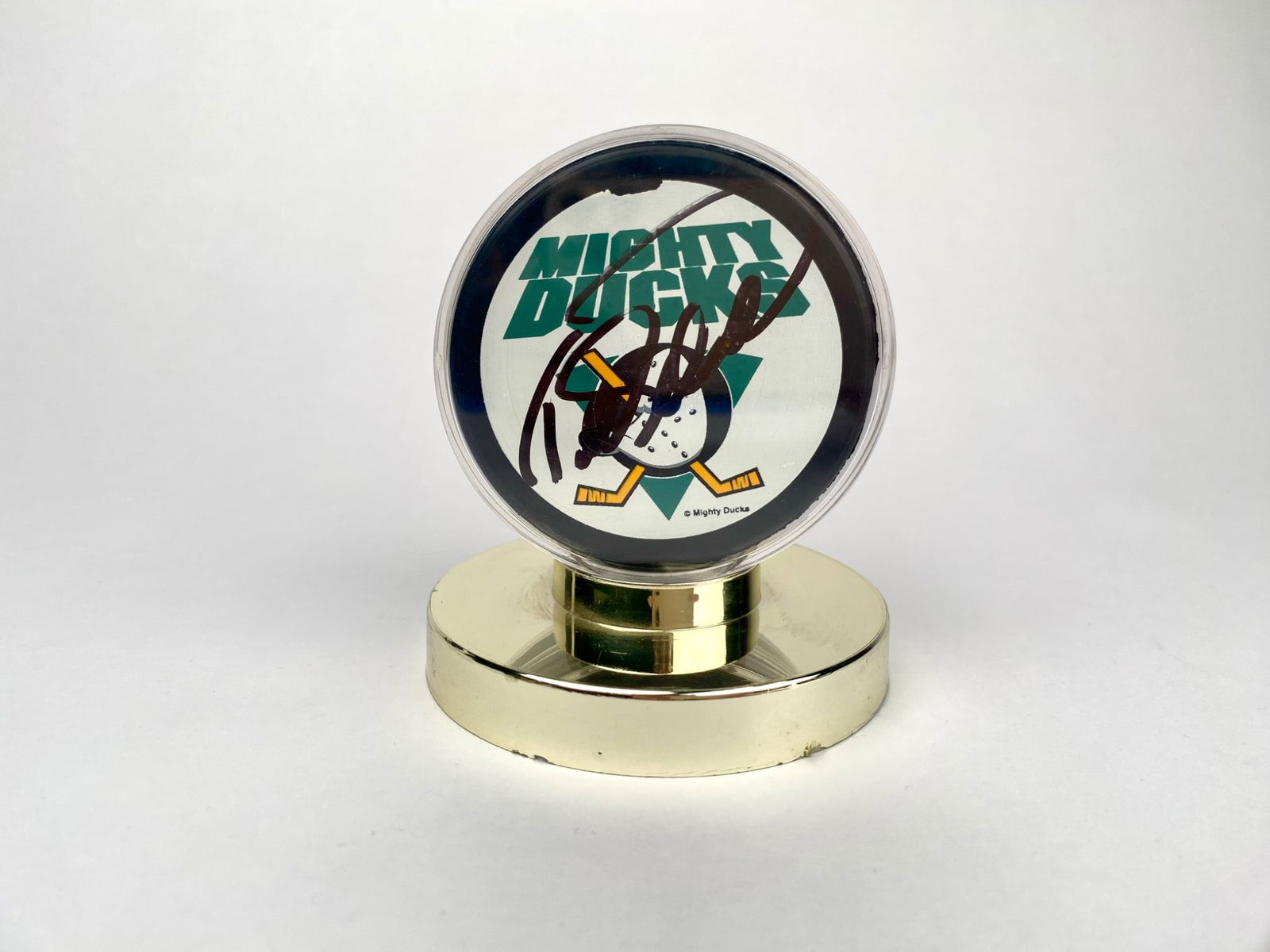 Todd Ewen Autographed Mighty Ducks Hockey Puck: Mighty Ducks collectible hockey puck, featuring the team’s logo and an autograph on the front from Todd Ewen. The hockey puck is housed in a clear protective case, mounted on a circular gold-tone ba