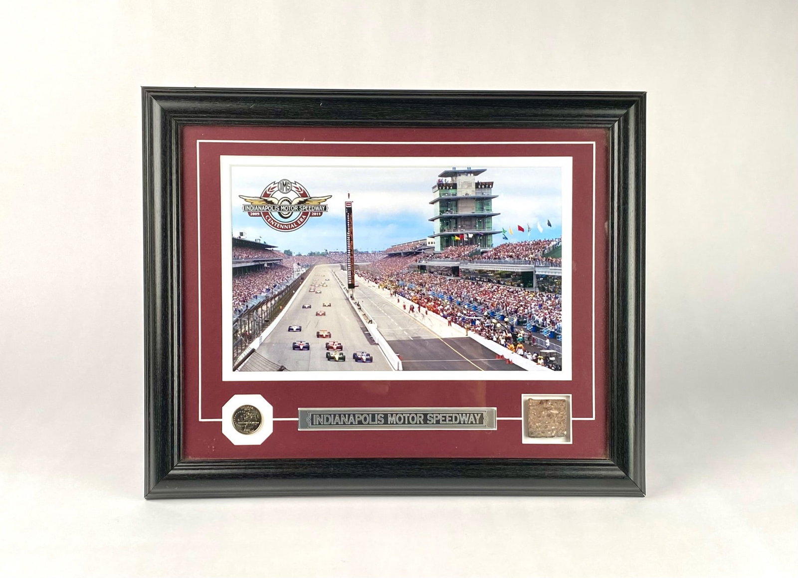 Indianapolis Motor Speedway Framed Display with Coin and Pavement Brick: Framed collectible display featuring an image of the Indianapolis Motor Speedway, accompanied by a commemorative coin and a small piece of pavement brick from the Speedway. The display includes a blac
