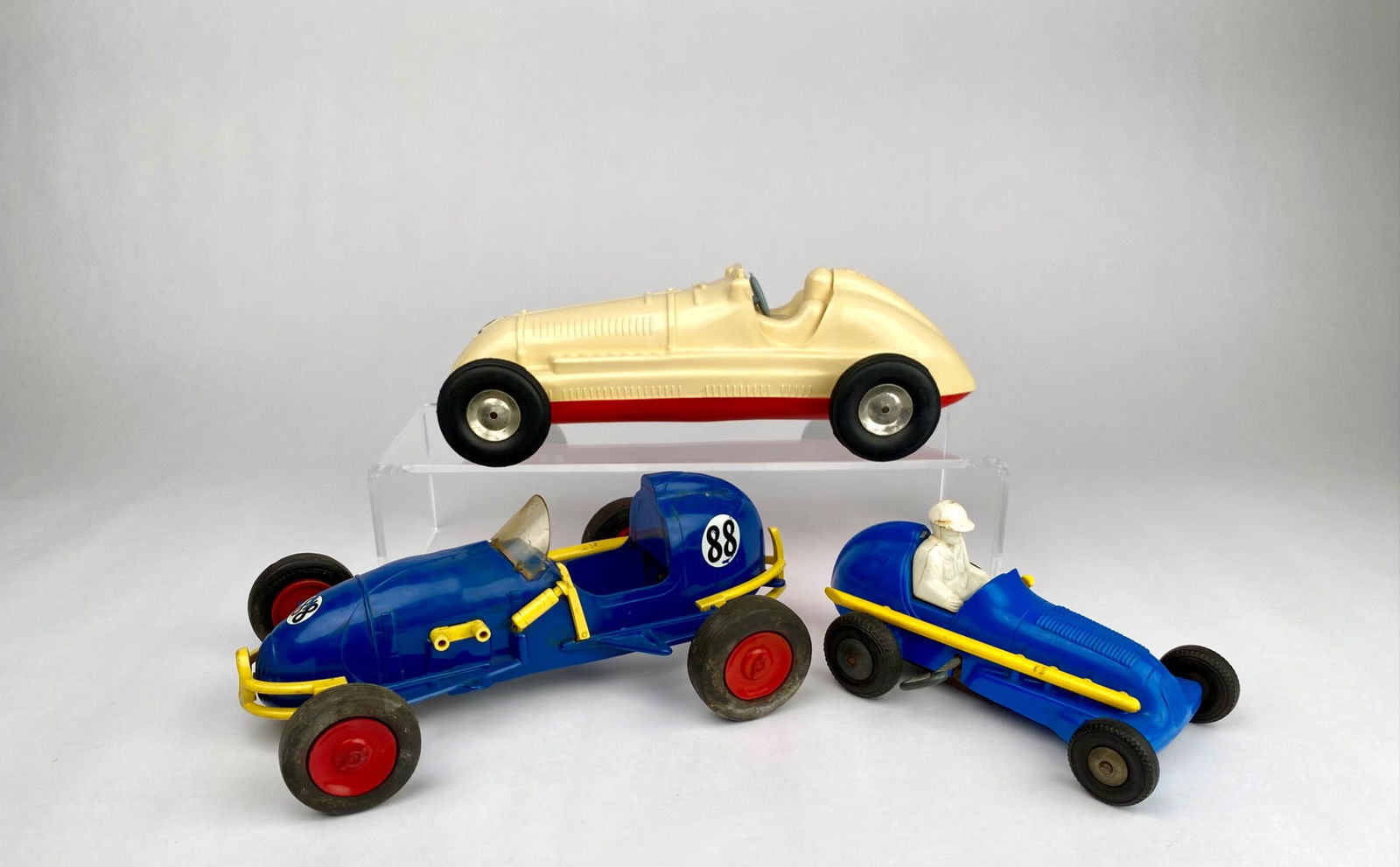 Three Vintage Plastic Racing Cars, Marx and Victory Industries: Three vintage plastic racing cars, including a cream-colored molded car with red accents and two blue racing cars featuring wind-up gears. Wear is visible on all three, commensurate with age and use.