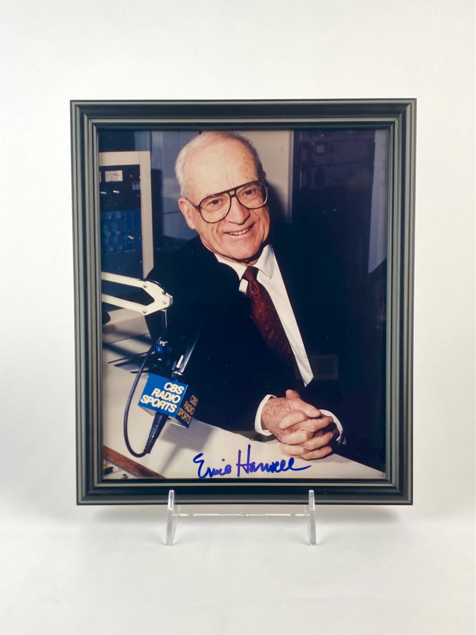 Framed Ernie Harwell Signed Photo: Framed color photograph of legendary broadcaster Ernie Harwell. The image depicts Harwell seated at a desk with a CBS Radio Sports microphone visible. The photo is signed in bold blue marker across th