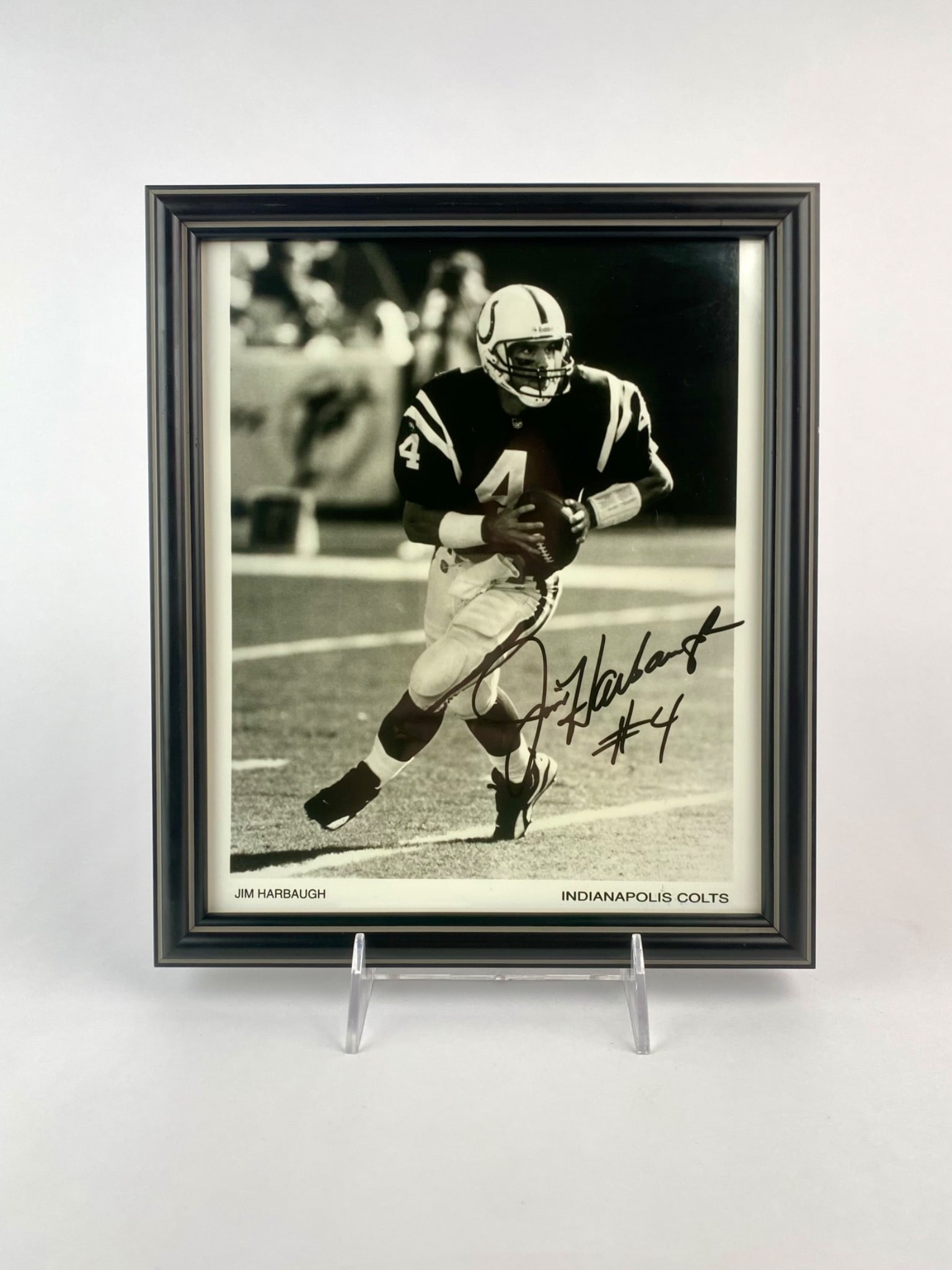 Jim Harbaugh Signed Indianapolis Colts Framed Photo: Black and white photograph of former NFL quarterback Jim Harbaugh in his Indianapolis Colts uniform, hand-signed in black marker with his jersey number “#4.” The photo is professionally framed