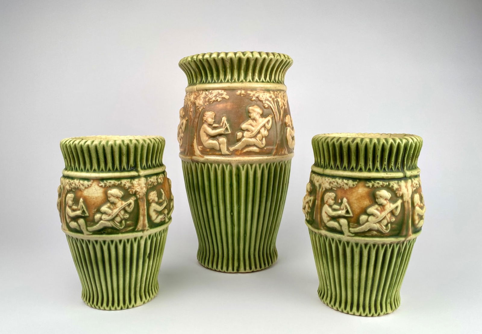 Roseville Pottery Donatello Three Piece Vase Lot, Early 20th Century: Three ceramic vases featuring green and beige tones with detailed relief scenes of children playing musical instruments in the Donatello pattern from Roseville Pottery. The design incorporates raised