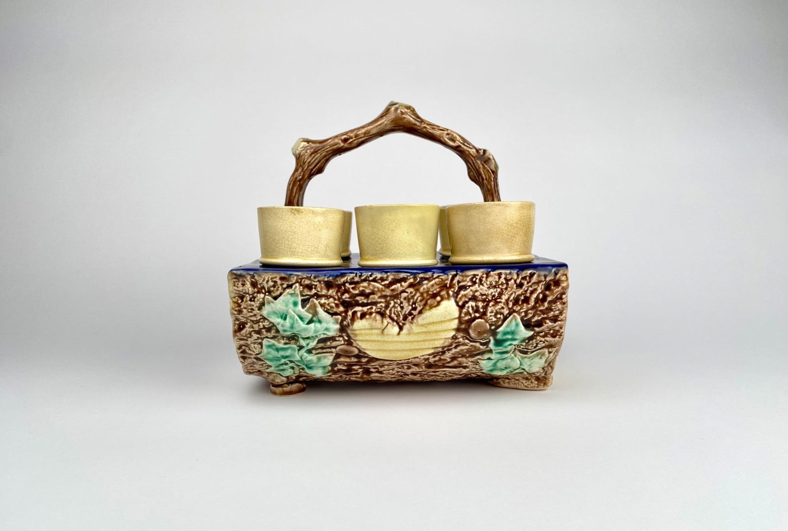 Majolica Egg Cup Holder with Six Cups: Majolica egg cup holder designed to resemble tree bark, featuring a molded branch-style handle and leaf details. Set includes six matching yellow-colored egg cups with a light pink interior. The holde