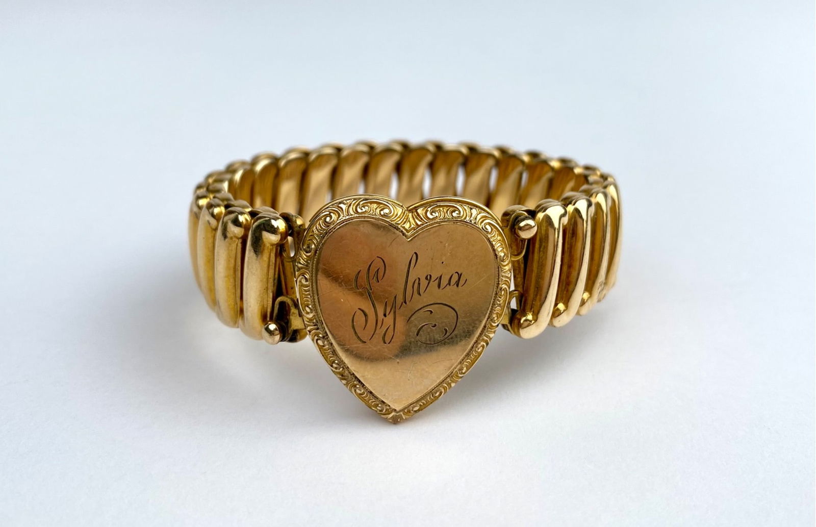 Vintage Sweetheart Expansion Bracelet with Heart Locket: Gold-tone vintage sweetheart expansion bracelet featuring a heart-shaped locket engraved with ‘Sylvia’ on the front. The locket opens to reveal two black and white photographs. The bracelet is mar