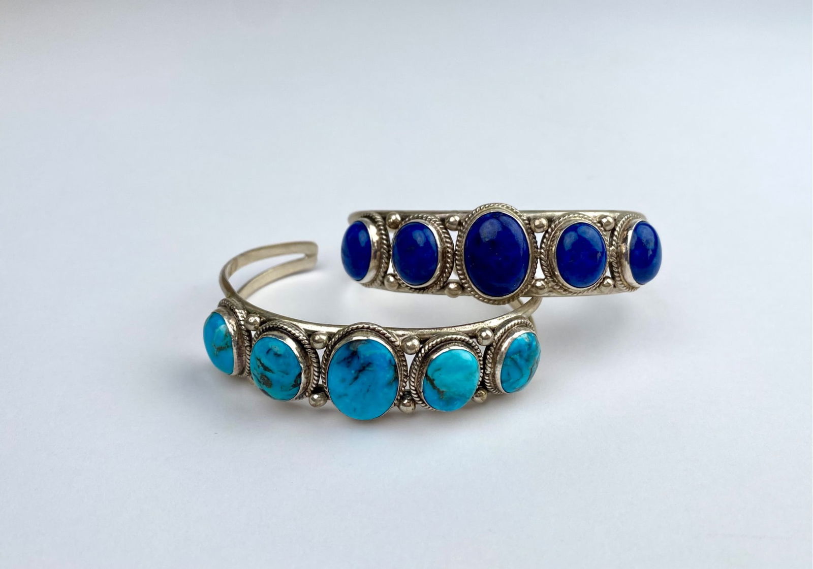 Pair of Sterling Silver Cuff Bracelets with Lapis Lazuli & Turquoise Stones: Sterling silver cuff bracelets featuring oval cabochon stones. One bracelet is set with vibrant blue lapis lazuli stones, and the other with turquoise stones. Both designs include detailed silverwork