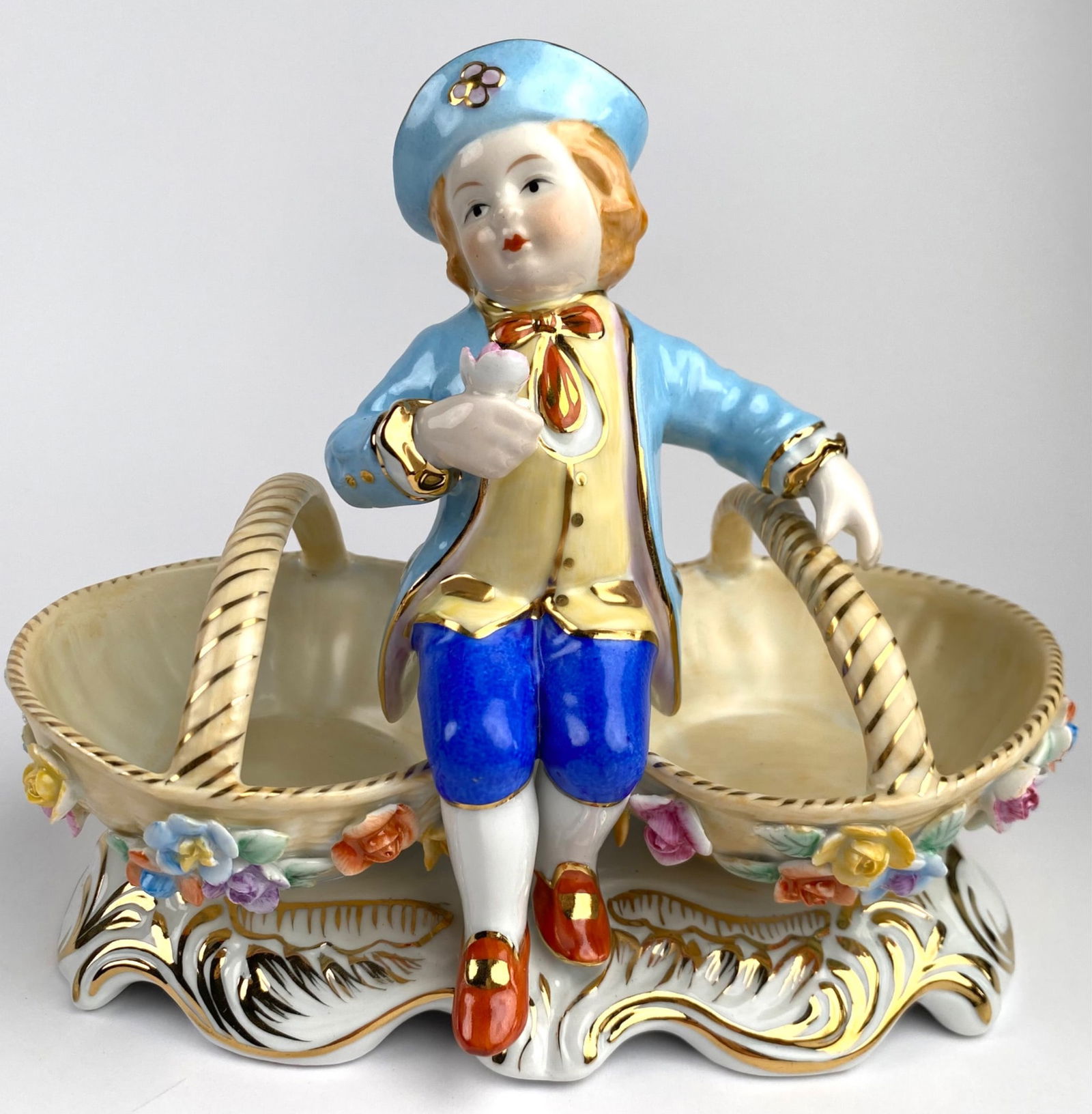 KPM Porcelain Boy Figurine with Dual Handled Basket and Floral Base: KPM porcelain figurine depicting a boy in a blue coat, pants, and hat, holding a pink flower, seated between dual-handled baskets. The baskets are adorned with multi-colored floral motifs and set on a
