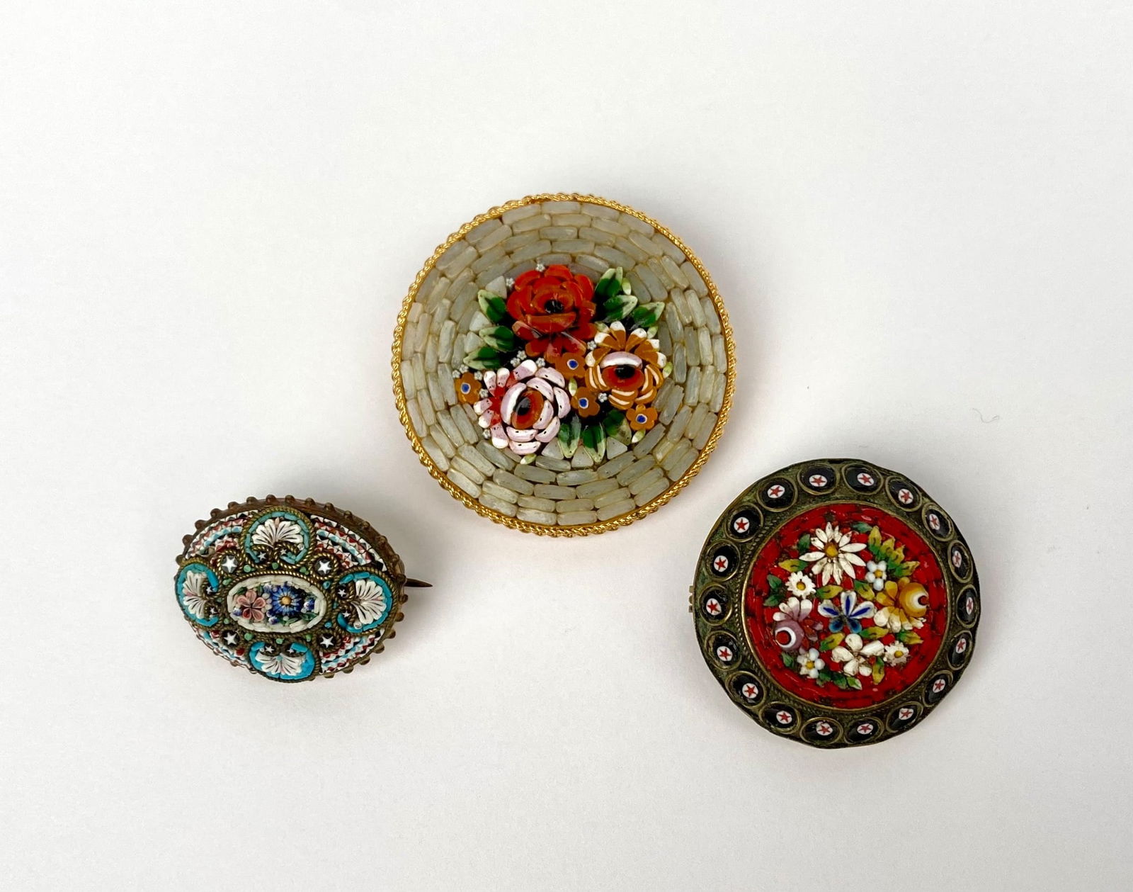 Three Italian Micro-Mosaic Floral Brooches: Three Italian micro-mosaic brooches, each showcasing floral designs. The largest brooch features a red rose centerpiece surrounded by a white and gray mosaic pattern, framed with gold-tone metal. The