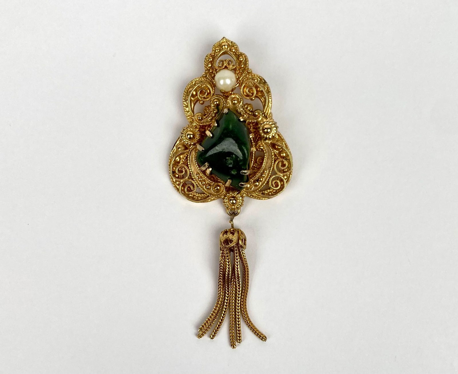 Gold-Tone Filigree Brooch With Green Stone, Pearl, & Tassel Detail: Gold-tone brooch featuring intricate filigree detailing, a central green stone, and a single pearl embellishment at the top. The piece is finished with a tassel detail consisting of multiple gold chai