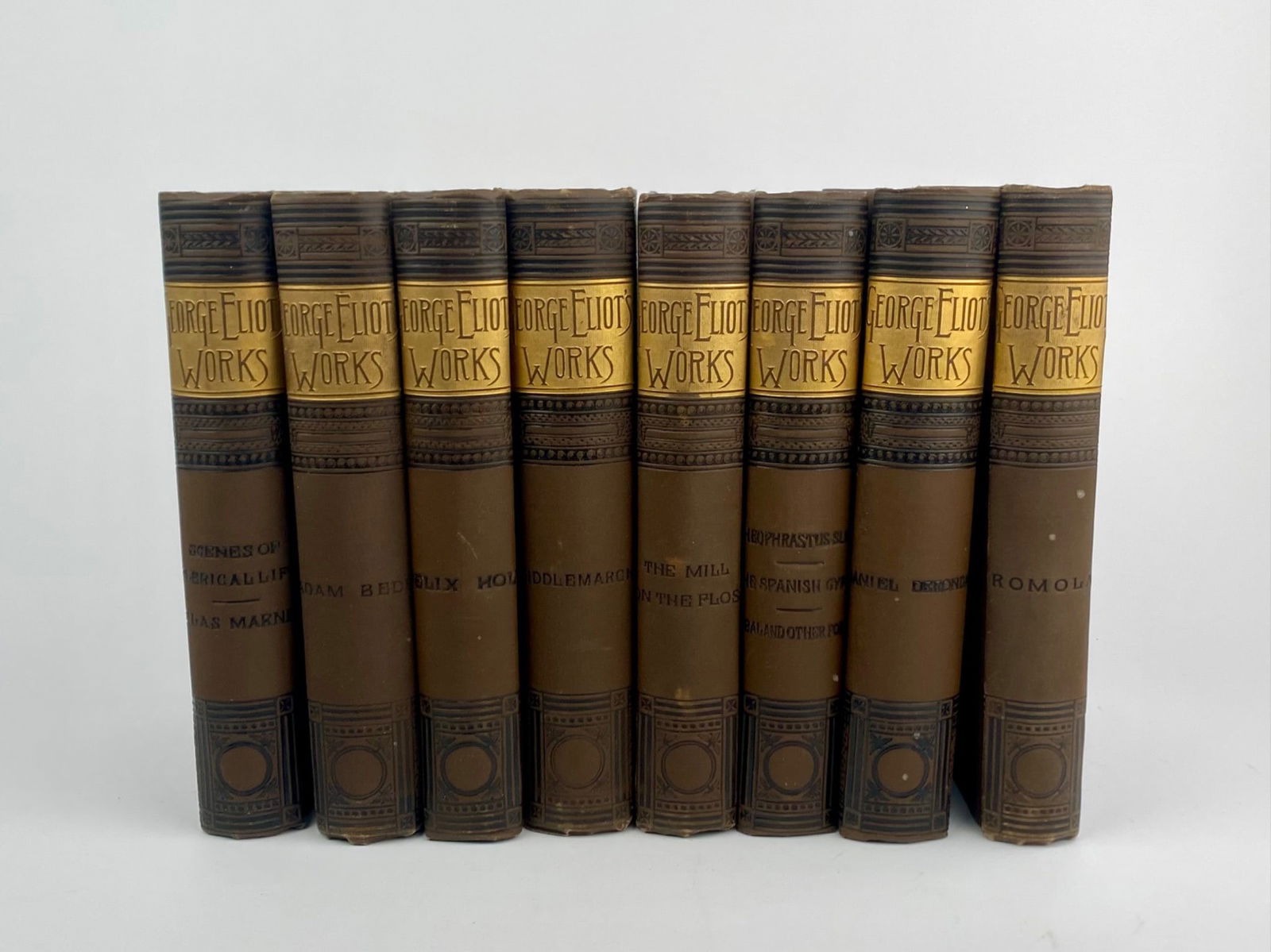 Eight Volume ‘George Eliot Works’ Decorative Hardcover Book Set: Eight volume hardcover set of ‘George Eliot Works,’ published by Hurst & Company, New York. The books are uniformly bound in brown cloth with decorative black stamping and gilt spine panels, and i